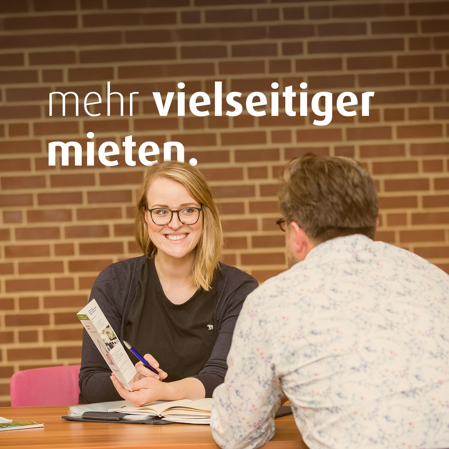 Eine Frau mit Brille und ein Mann sitzen sich gegenüber am Tisch und unterhalten sich. Die Frau zeigt dem Mann eine Broschüre. Auf dem Bild steht der Text "mehr vielseitiger mieten".