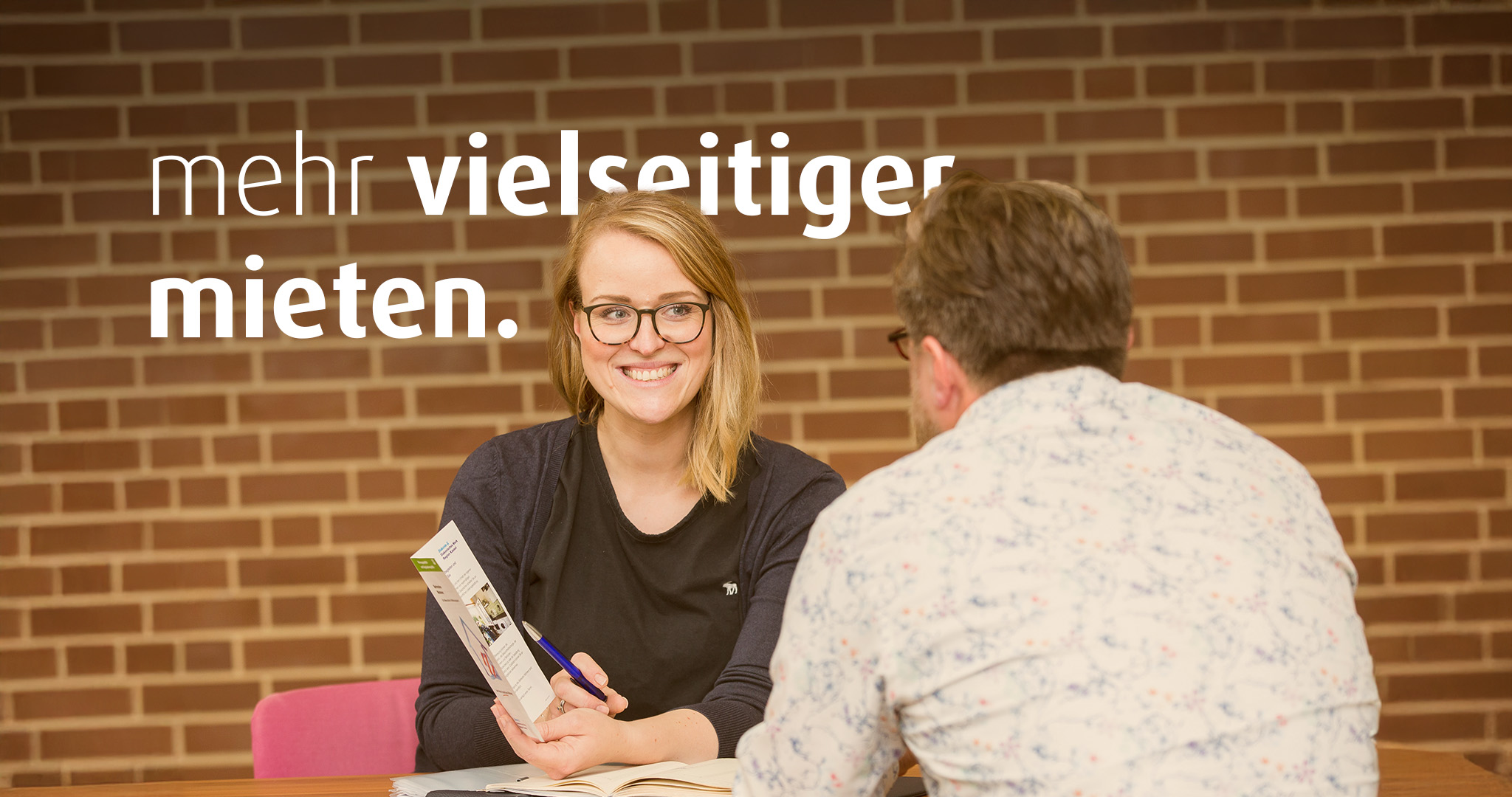 Eine Frau mit Brille und ein Mann sitzen sich gegenüber am Tisch und unterhalten sich. Die Frau zeigt dem Mann eine Broschüre. Auf dem Bild steht der Text "mehr vielseitiger mieten".