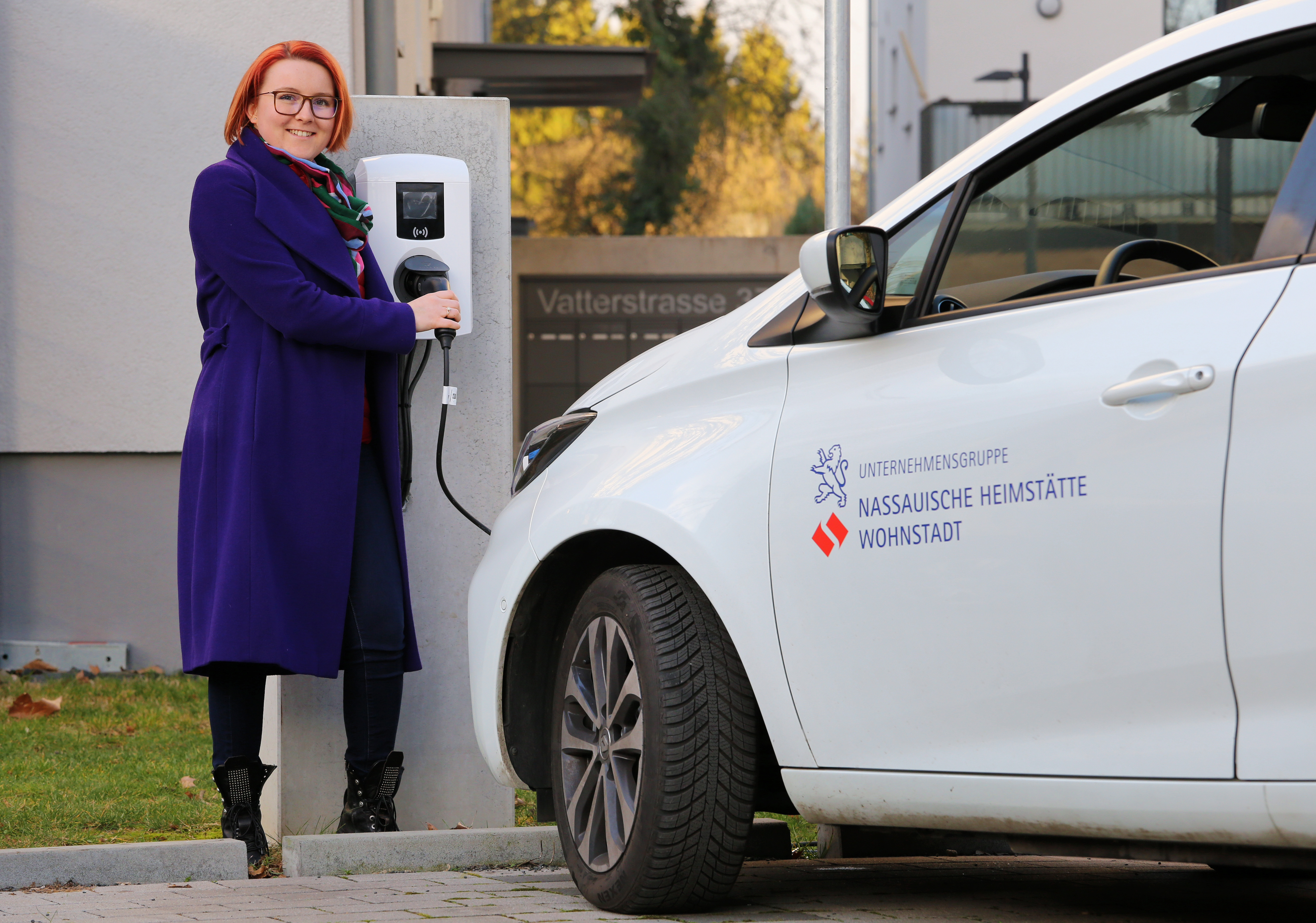 Frau steht an einer Elektrotanksäule und hält den Stecker in der Hand. Davor parkt ein weißes Elektroauto.