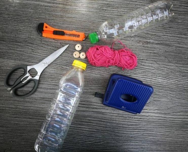 PET-Flaschen, Messer, Schere, Wolle, Locher und Holzperlen liegen auf einem Tisch.