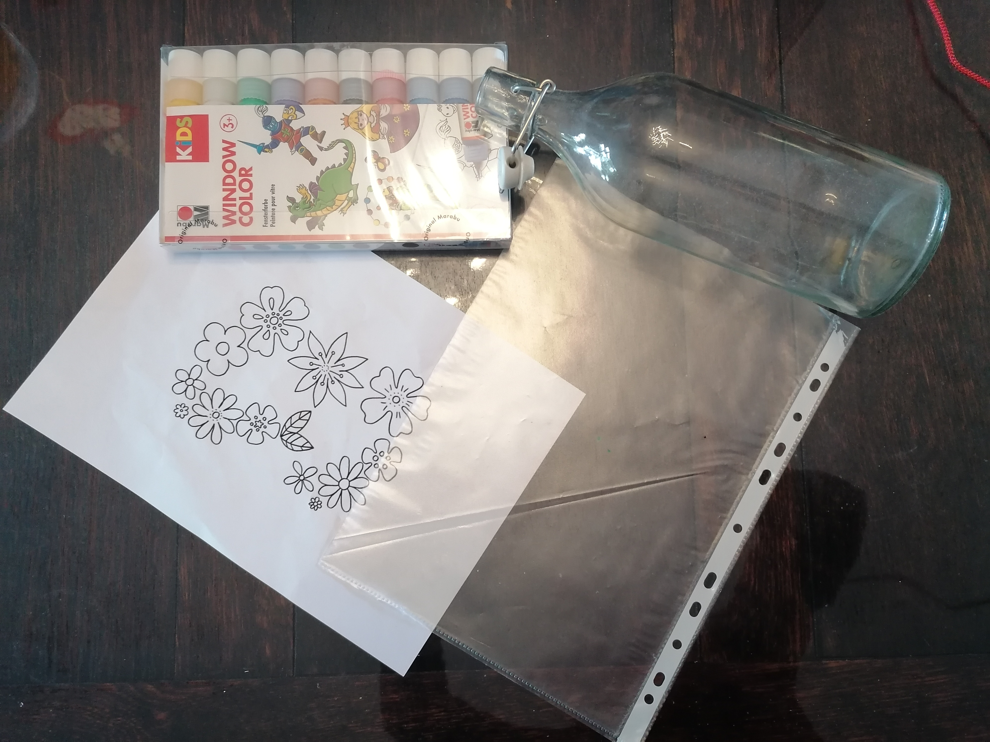 Glasflache, Folie, Windowcolor und Malvorlage für Blumen liegen auf einem Tisch.