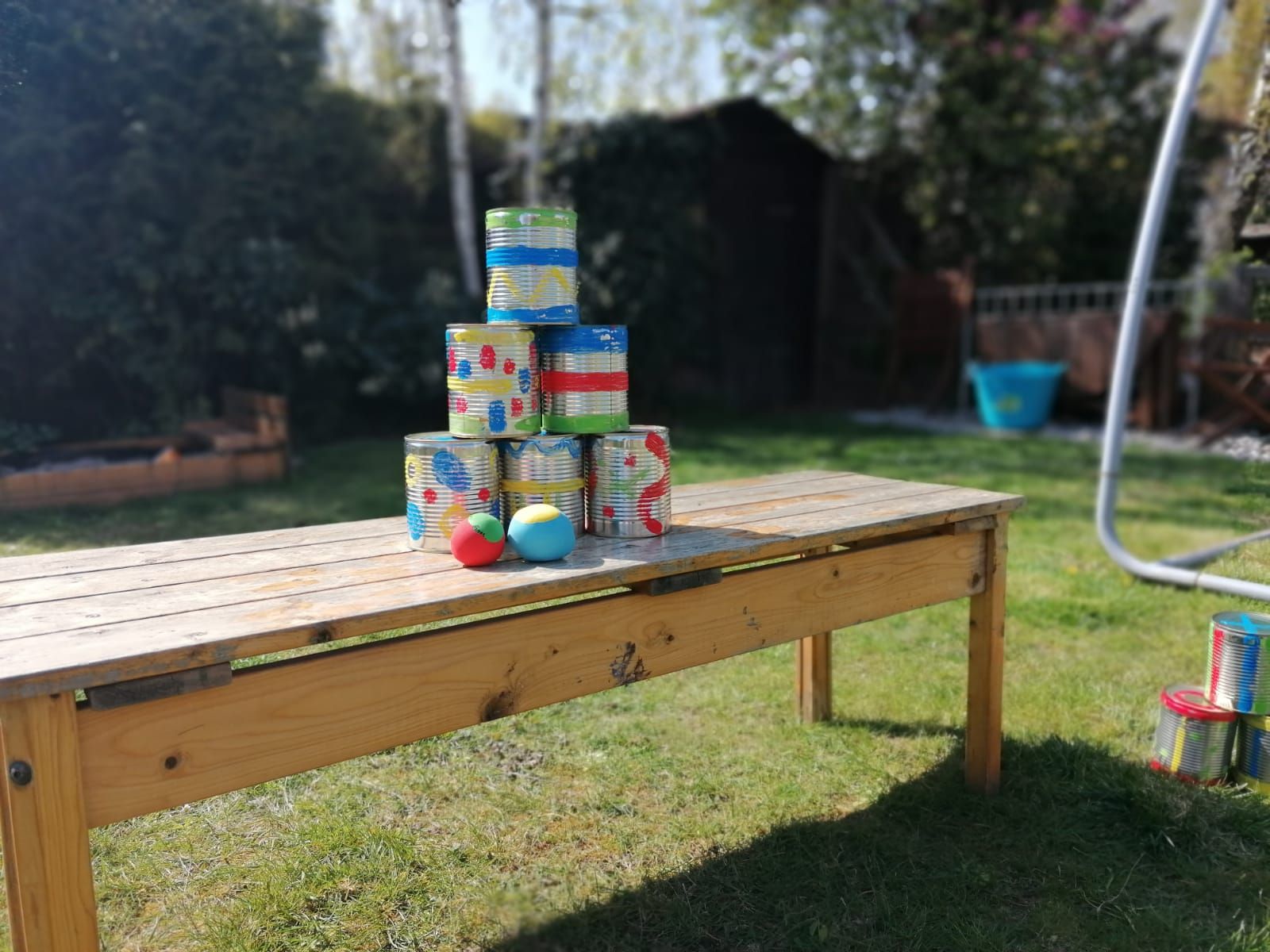 Im Garten steht ein Tisch. Auf dem Tisch stehen bunte Dosen. Sie sind zu einer Pyramide aufgebaut.