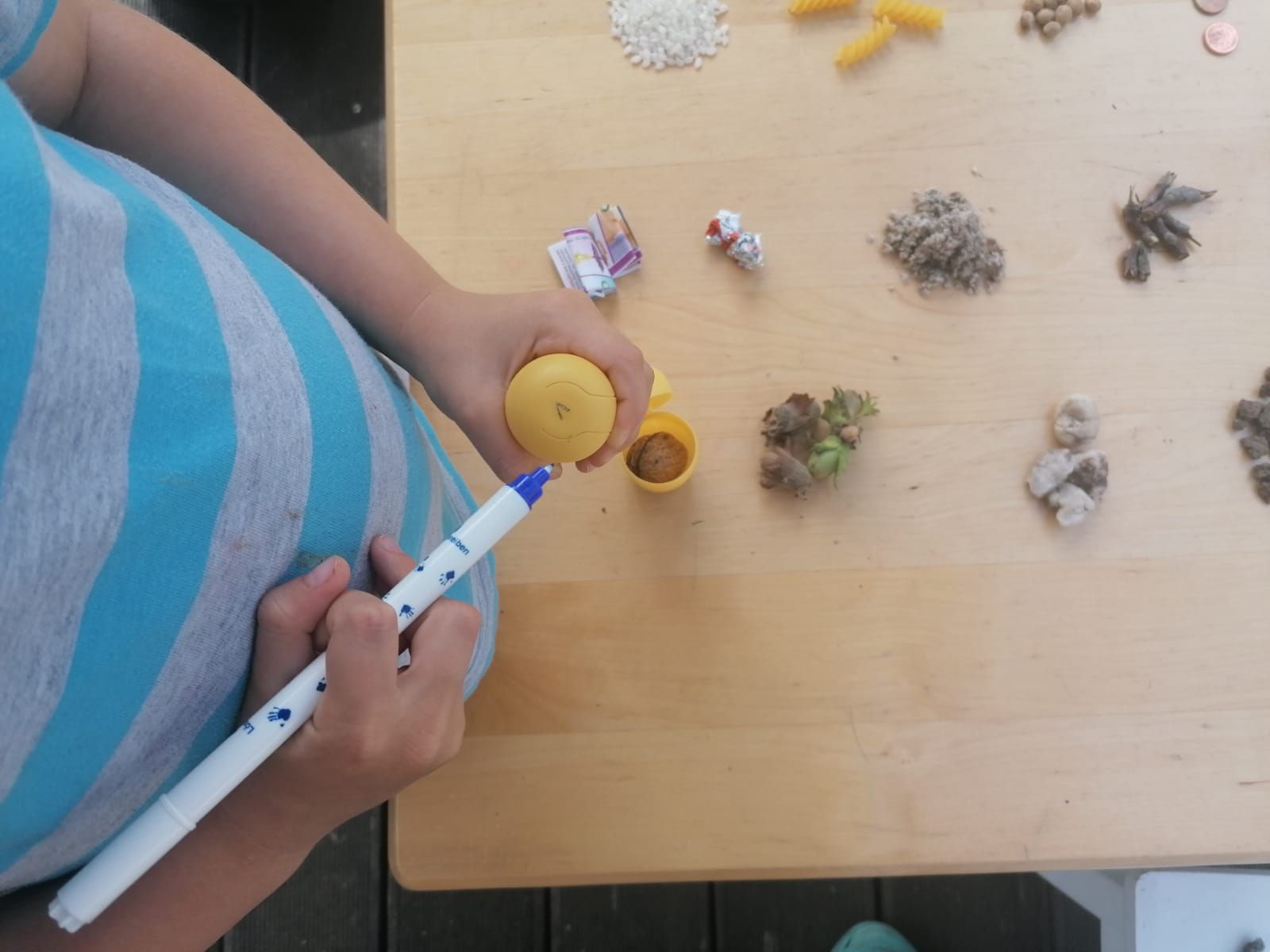 Junge beschriftet eine kleine, gelbe Verpackung. Auf dem Tisch liegen kleine Mengen von Reis, Nudeln, Erbsen, Steine etc.