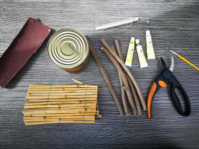 Konservendose, Schmirgelpapier, Farbe, Pinsel, Klarlack, Draht, Stäbe, Äste und Gartenschere liegen auf einem Tisch.