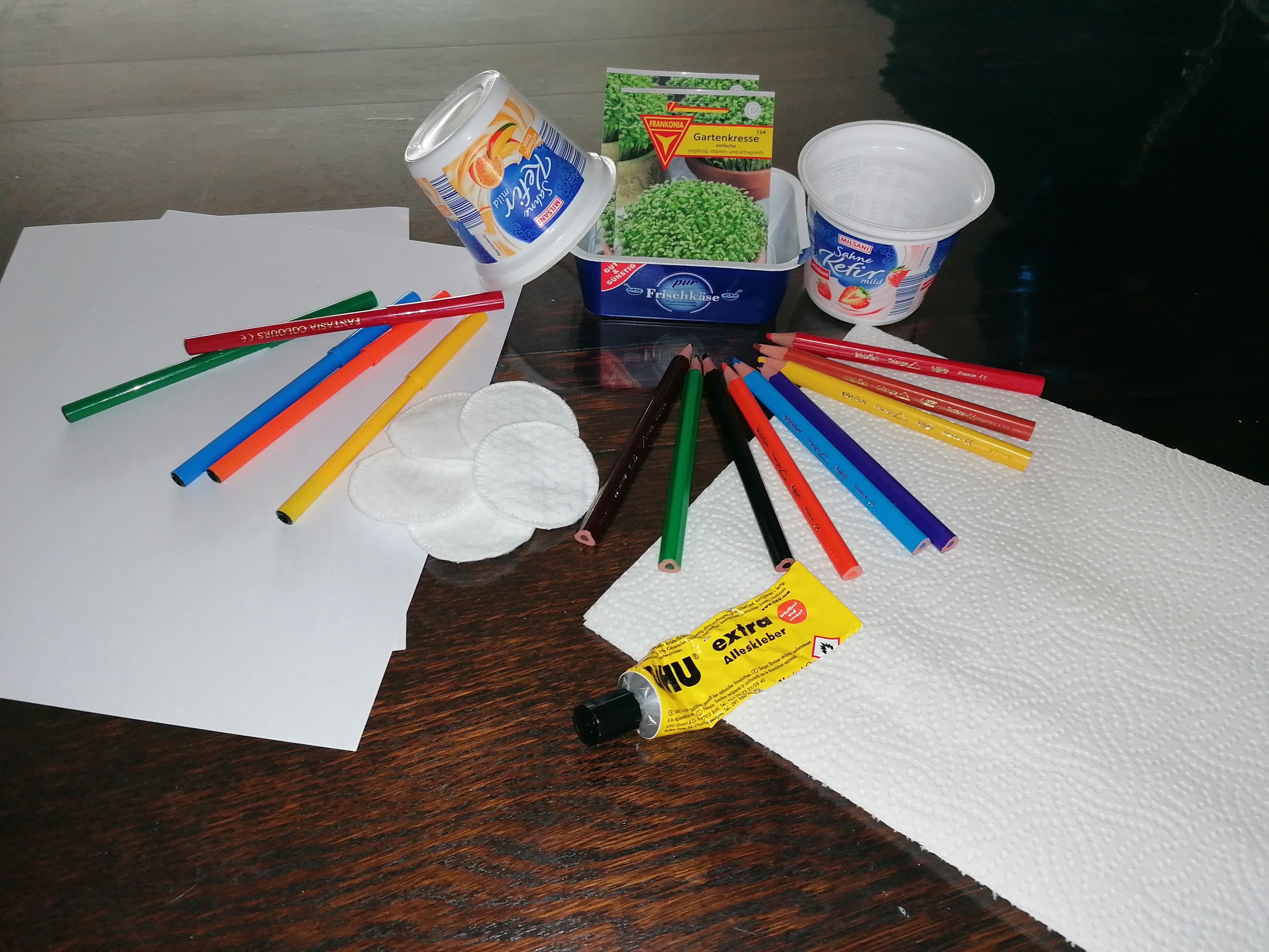 Joghurtbecher, Kressesamen, Watte, Blätter, Stifte und Kleber liegen auf einem Tisch.