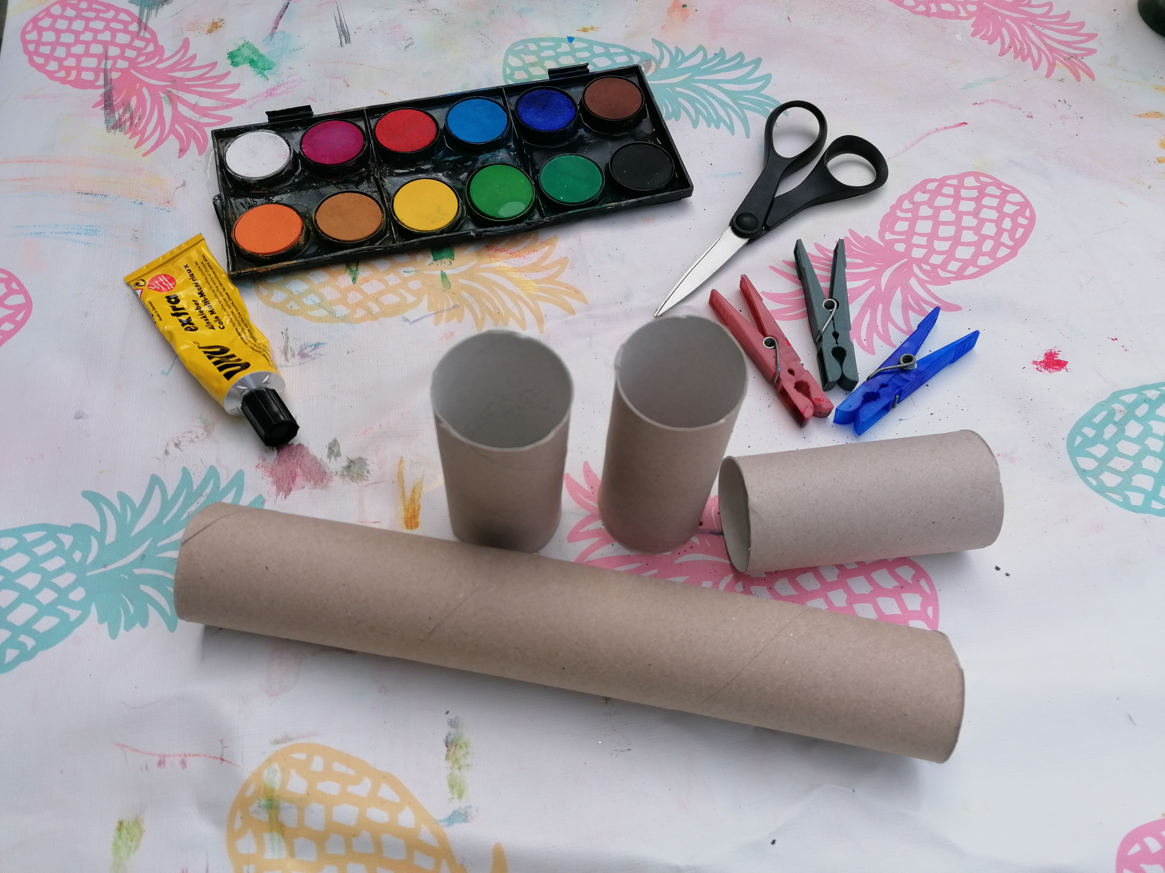 Papierrollen, Wäscheklammern, Schere, Kleber und Wasserfarbkasten liegen auf einem Tisch.