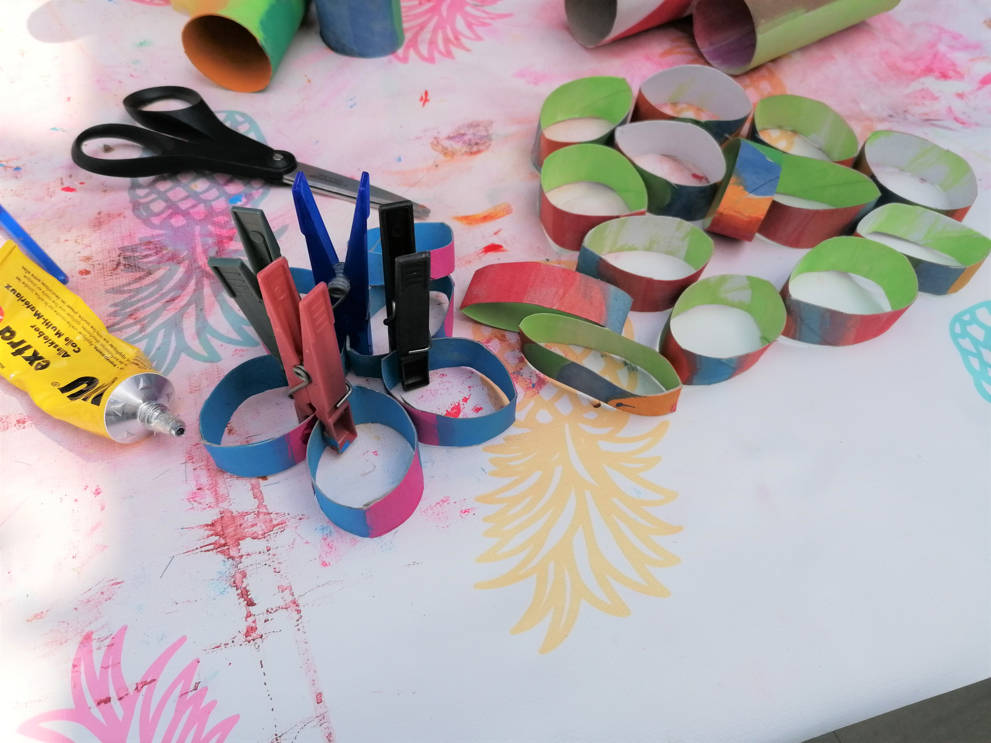 Angemalte und in Streifen geschnittene Papierrollen, Kleber, Schere und Wäscheklammern auf einem Tisch.