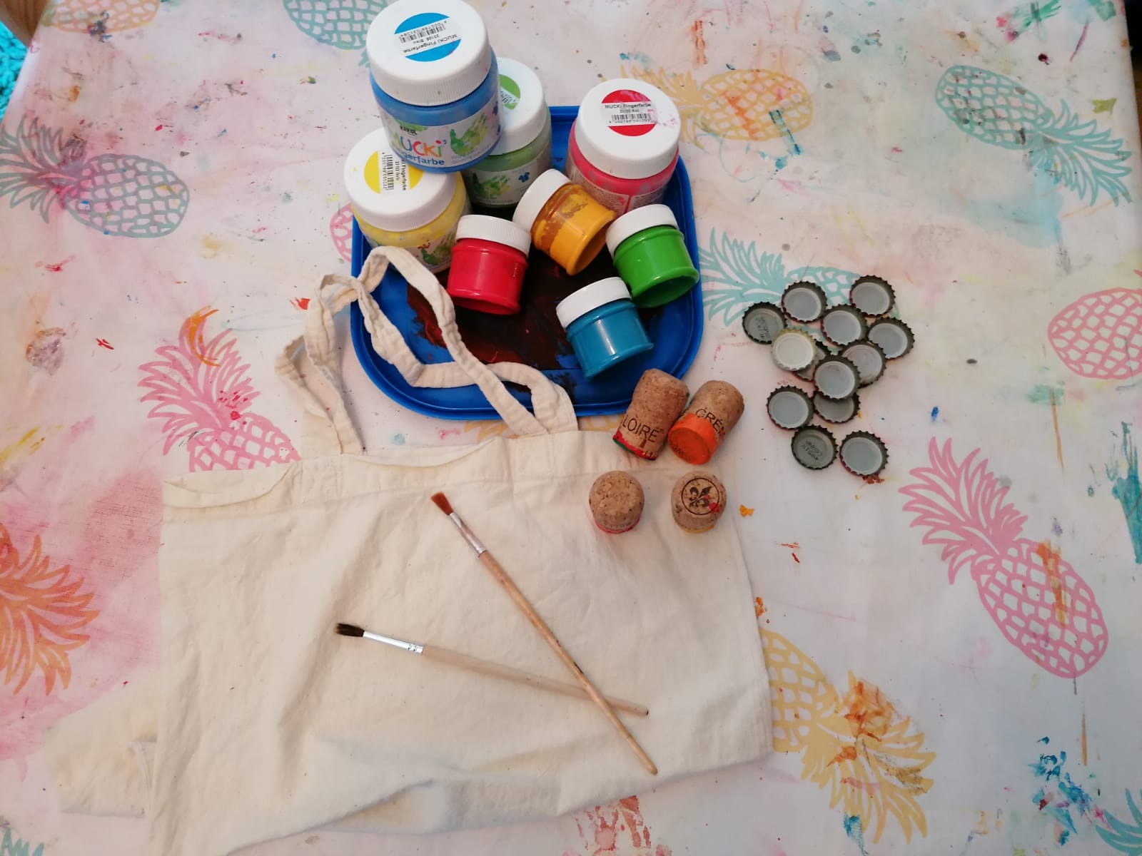 Stoffbeutel, Pinsel, Korken, Kronkorken und Farbe liegen auf einem Tisch.