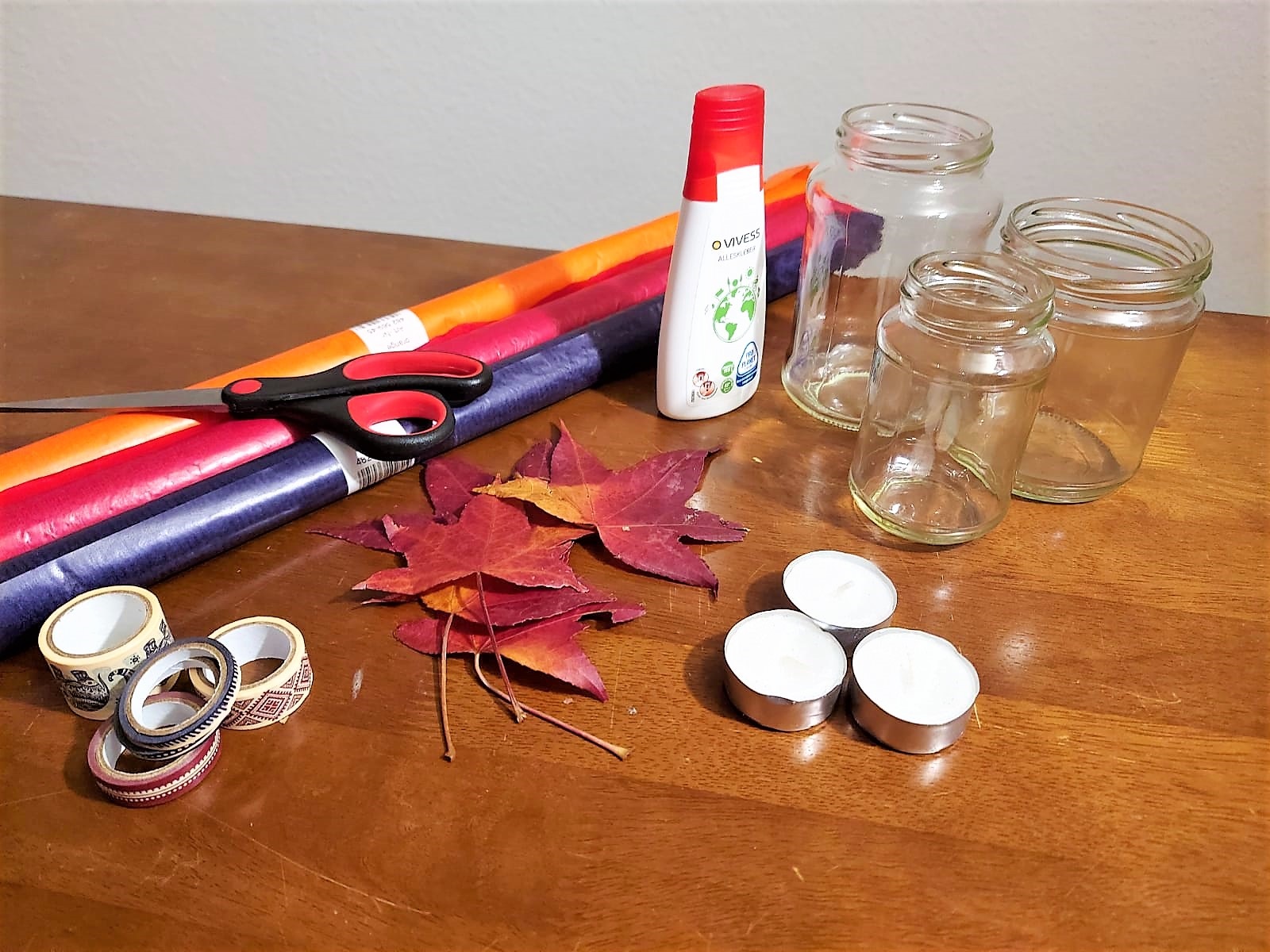 Einweggläser, Kleber, Teelichter, Schere, Kleber und Dekomaterial liegen auf dem Tisch.