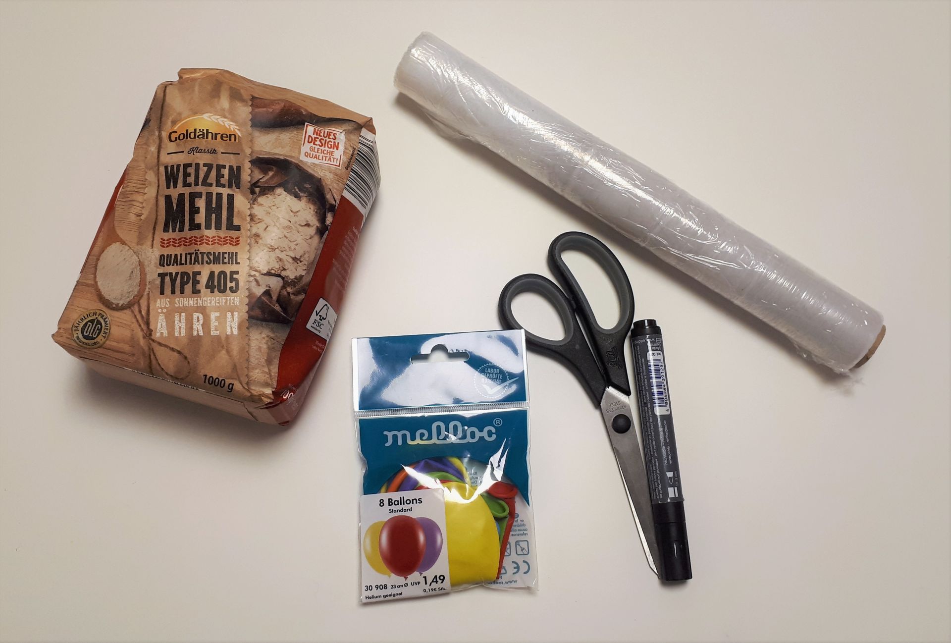 Frischhaltefolie, Luftballons, Mehl, Schere und wasserfester Filzstift liegen auf einem Tisch.