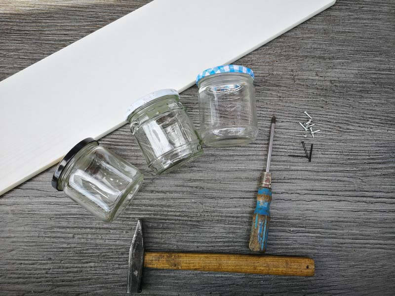 Drei leere Marmeladengläser mit Deckel, ein Holzbrett, Schrauben, Schraubenzieher, Hammer und Nägel liegen auf einem Tisch.