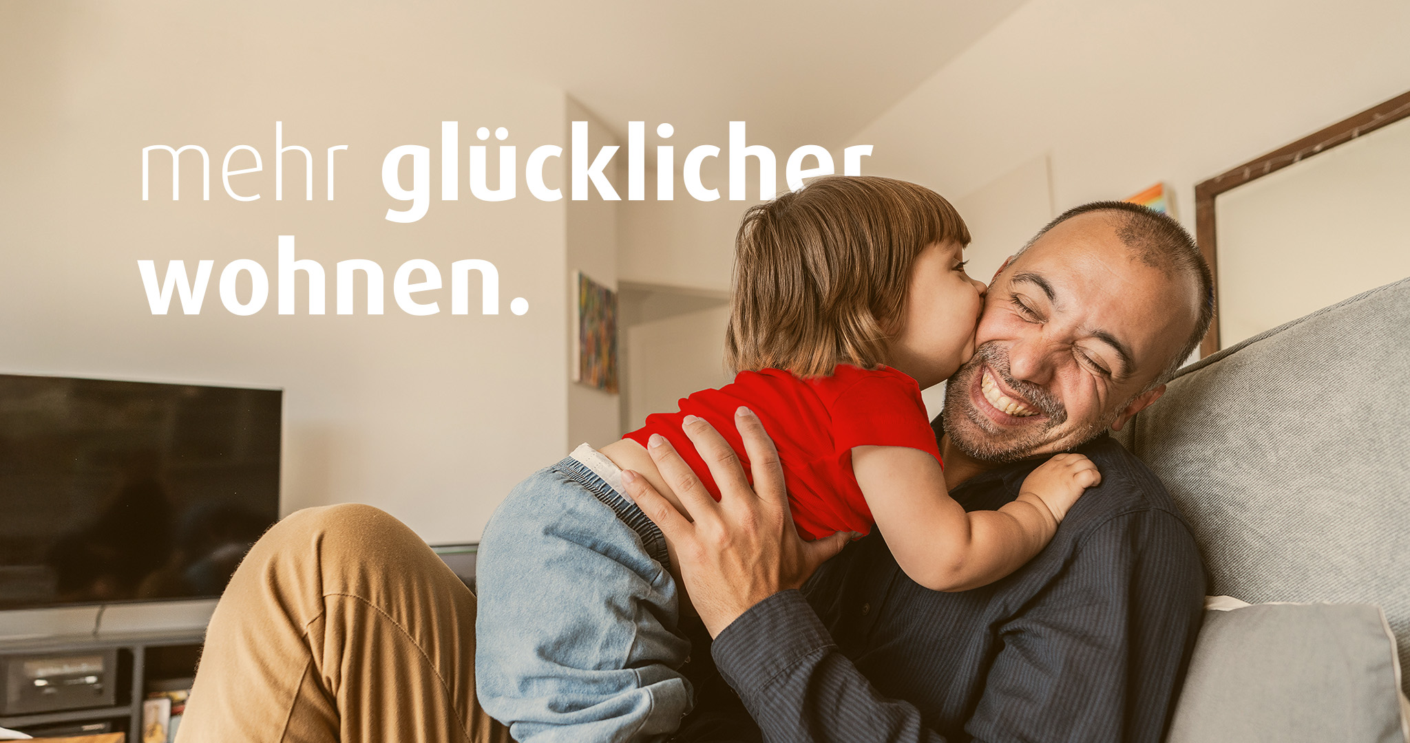 Ein Mann sitzt lächelnd auf dem Sofa und hat ein kleines Kind mit rotem Shirt auf dem Schoß. Das Kind gibt dem Mann einen Kuss auf die Wange. Auf dem Bild steht "mehr glücklicher wohnen."