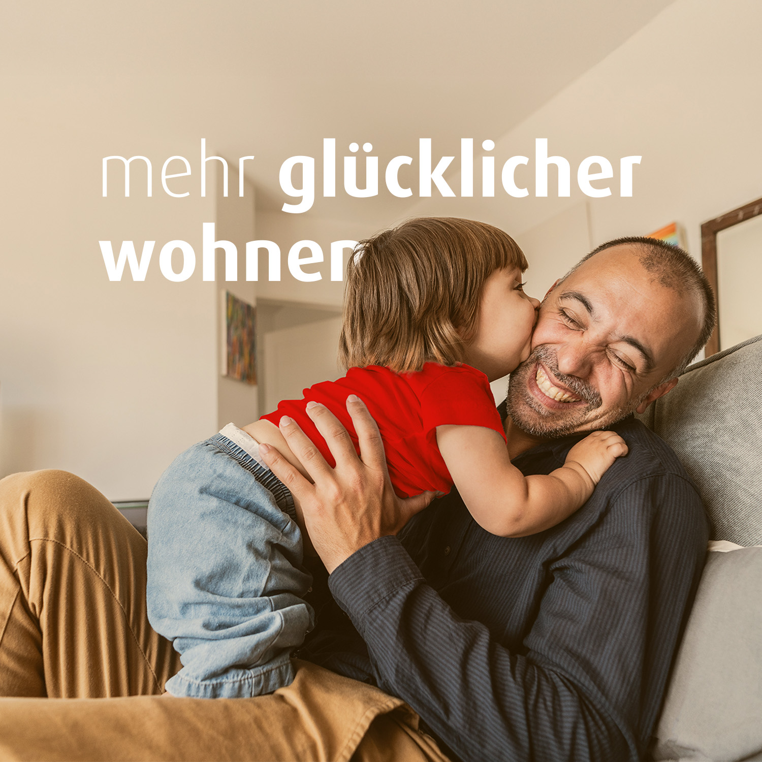 Ein Mann sitzt lächelnd auf dem Sofa und hat ein kleines Kind mit rotem Shirt auf dem Schoß. Das Kind gibt dem Mann einen Kuss auf die Wange. Auf dem Bild steht "mehr glücklicher wohnen."