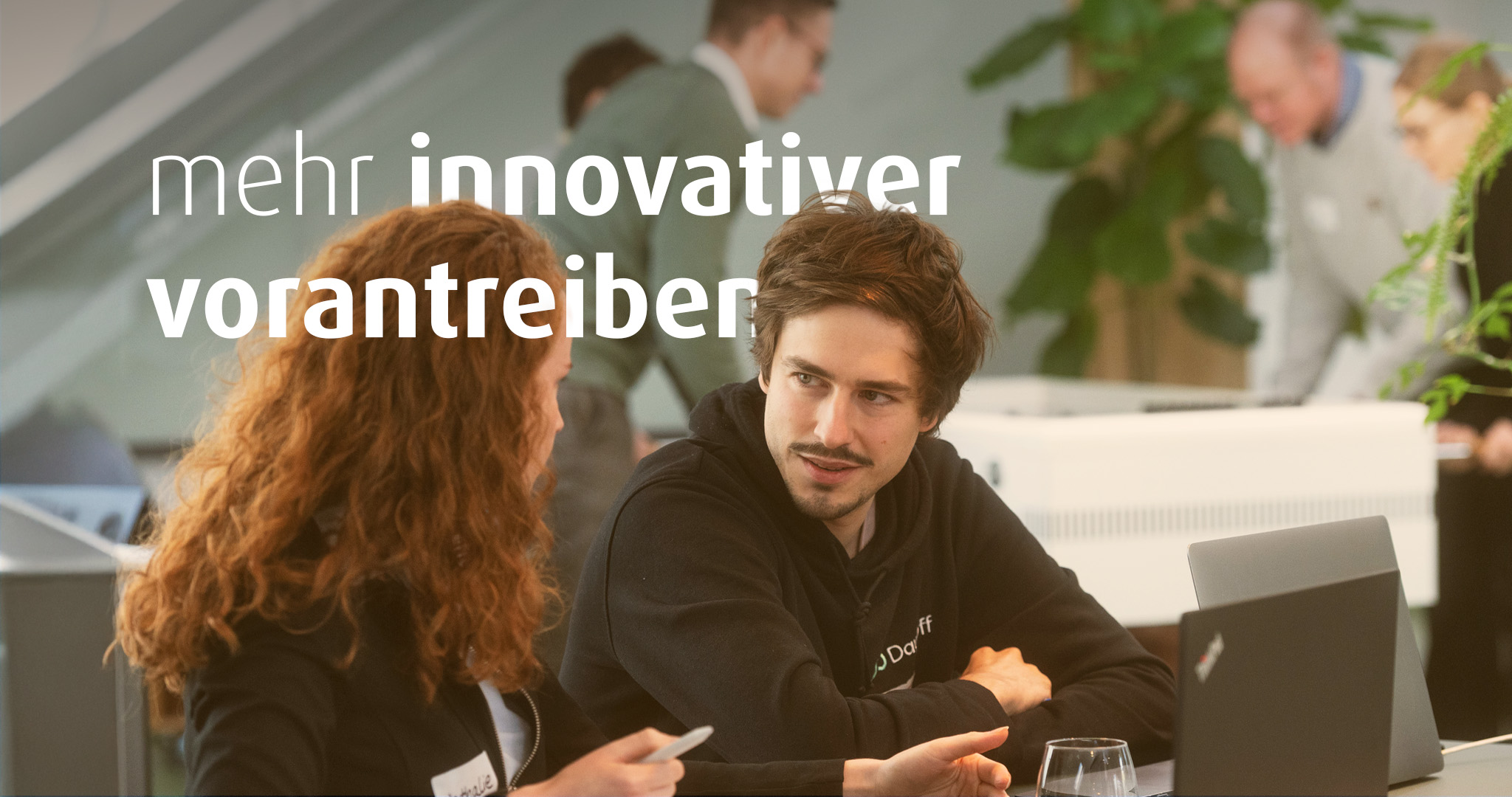 Eine Frau und ein Mann sitzen nebeneinander an einem Tisch. Vor ihnen stehen Laptops. Im Bild steht der Text "mehr innovativer vorantreiben".