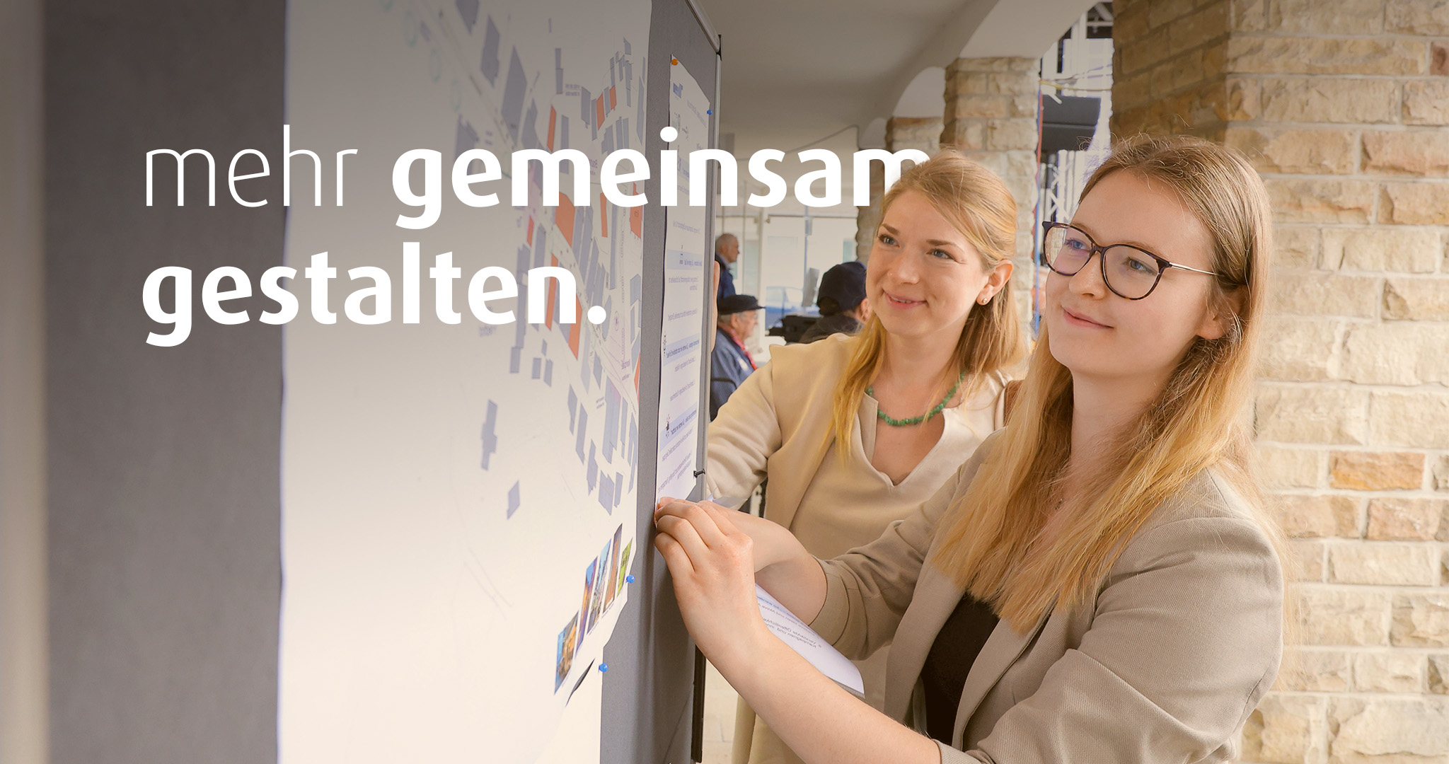 Zwei junge Frauen schauen auf einen Plan, der an der Wand befestigt ist. Auf dem Bild steht der Text "mehr gemeinsamer gestalten".