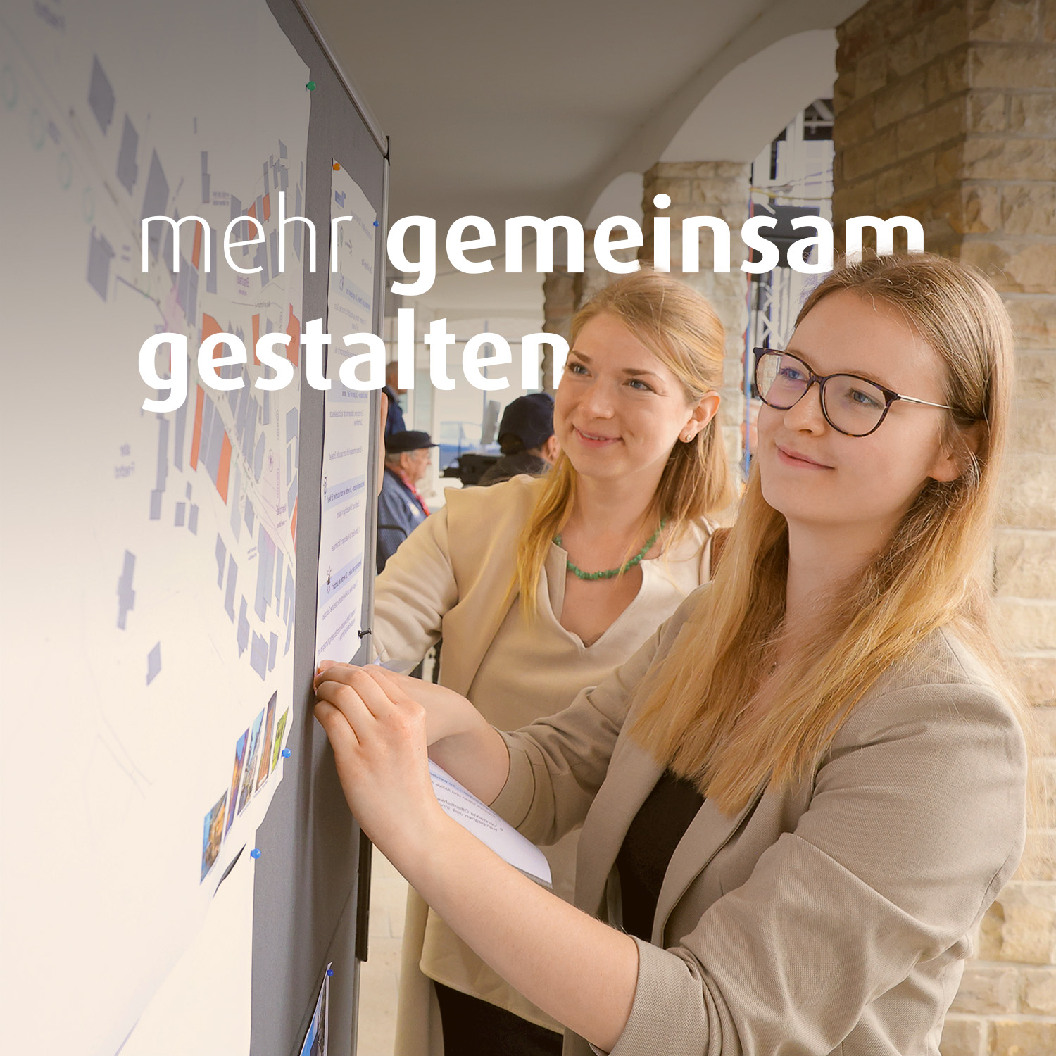 Zwei junge Frauen schauen auf einen Plan, der an der Wand befestigt ist. Auf dem Bild steht der Text "mehr gemeinsamer gestalten".
