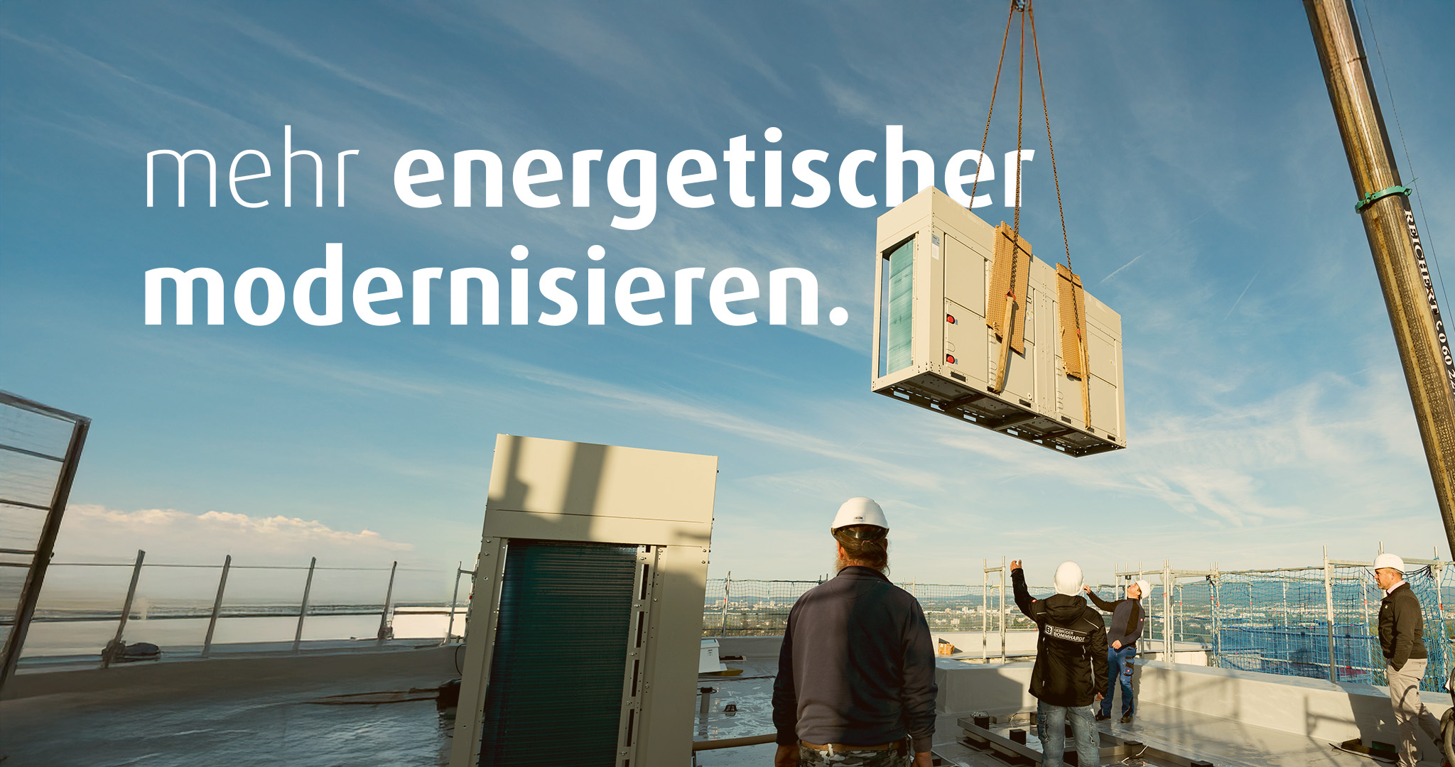 Mit einem Kran wird eine Wärmepumpe auf ein Hausdach gehoben. Vier Bauarbeiter stehen auf dem Dach. Auf dem Bild steht der Text "mehr energetischer modernisieren".