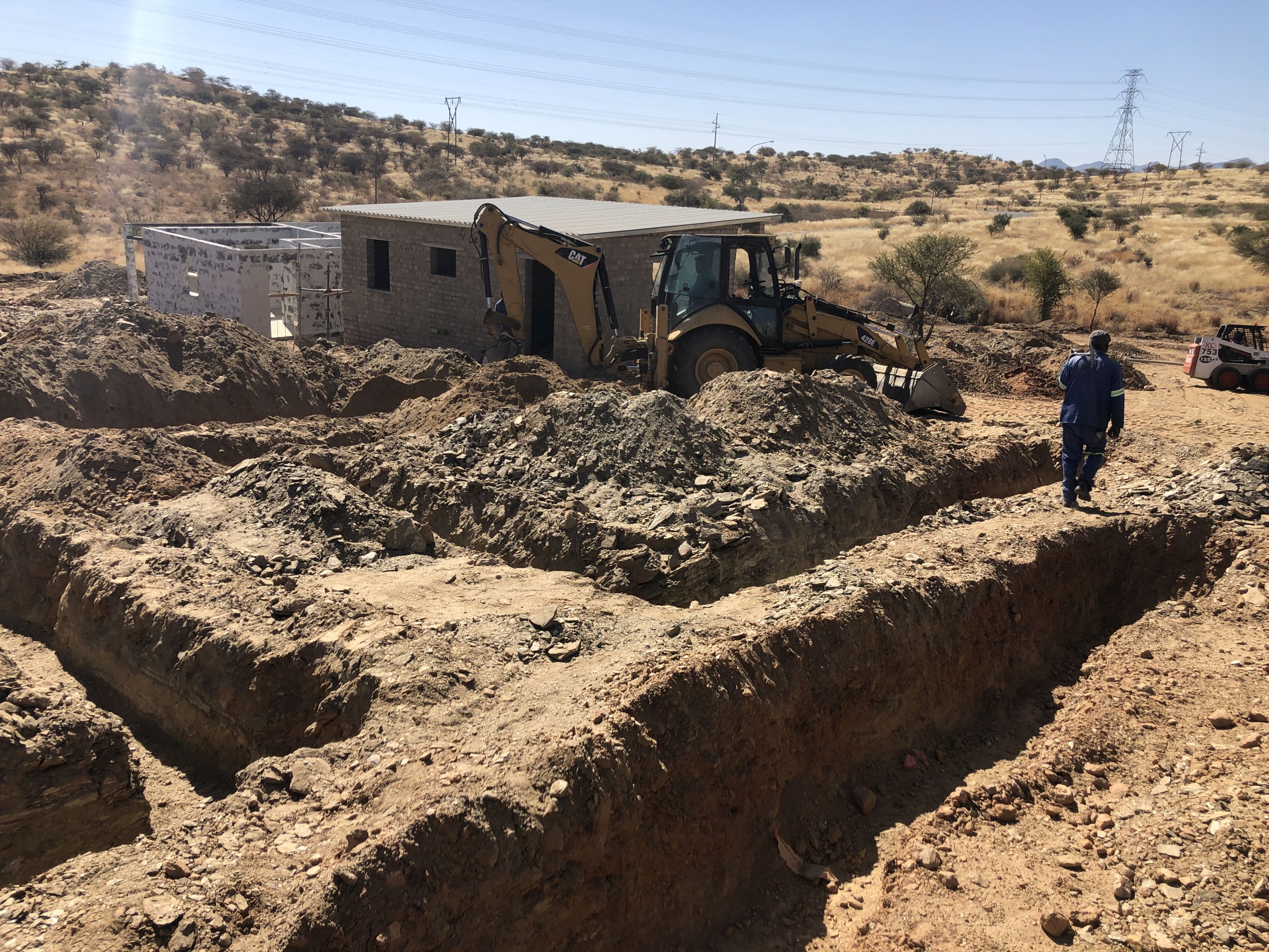 Ein Mann und ein Bagger auf einer Baustelle mit zwei Häusern, viel Sand und trockene Erde