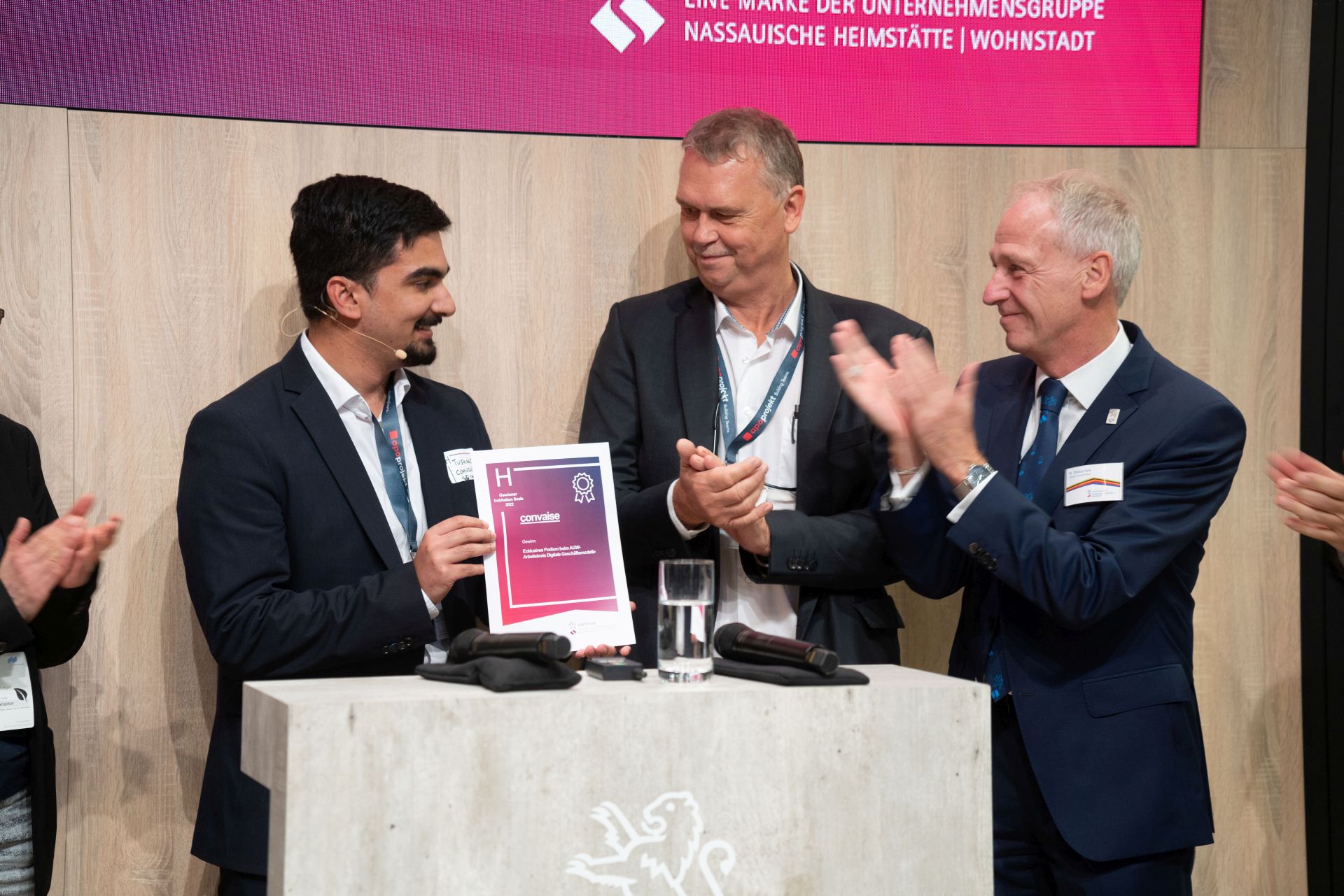 Drei Männer im Anzug, einer mit Urkunde, die anderen beiden applaudieren