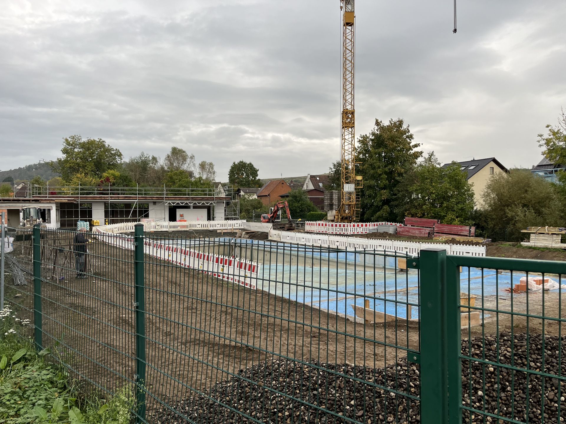 Blick auf das Freibad von Großen-Buseck mit Gerüst und Baustellenabsperrungen, im Vordergrund ein grüner Zaun, im Hintergrund ein Kran