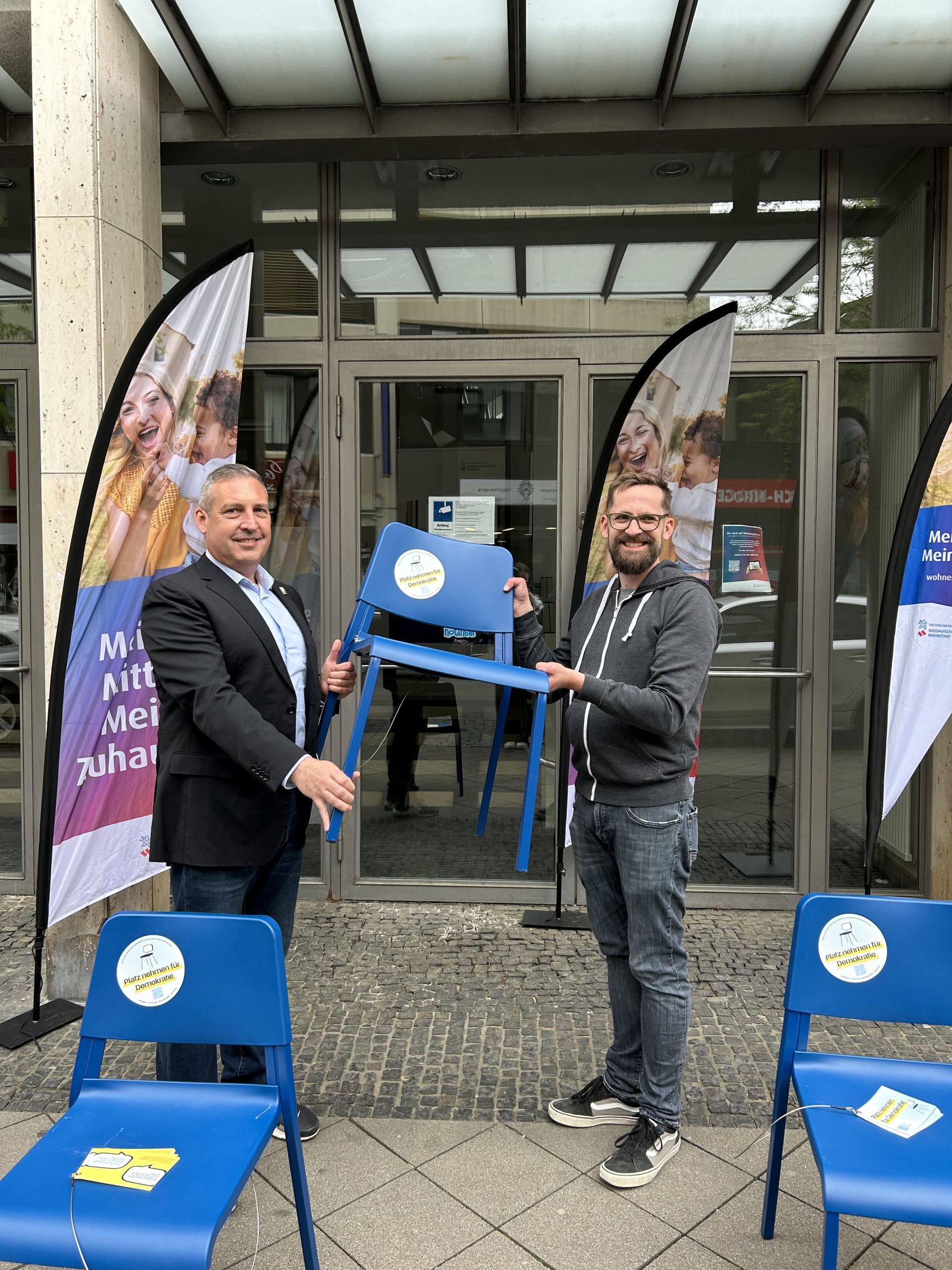 Zwei Männer halten einen blauen Stuhl, im Vordergrund zwei weitere blaue Stühle