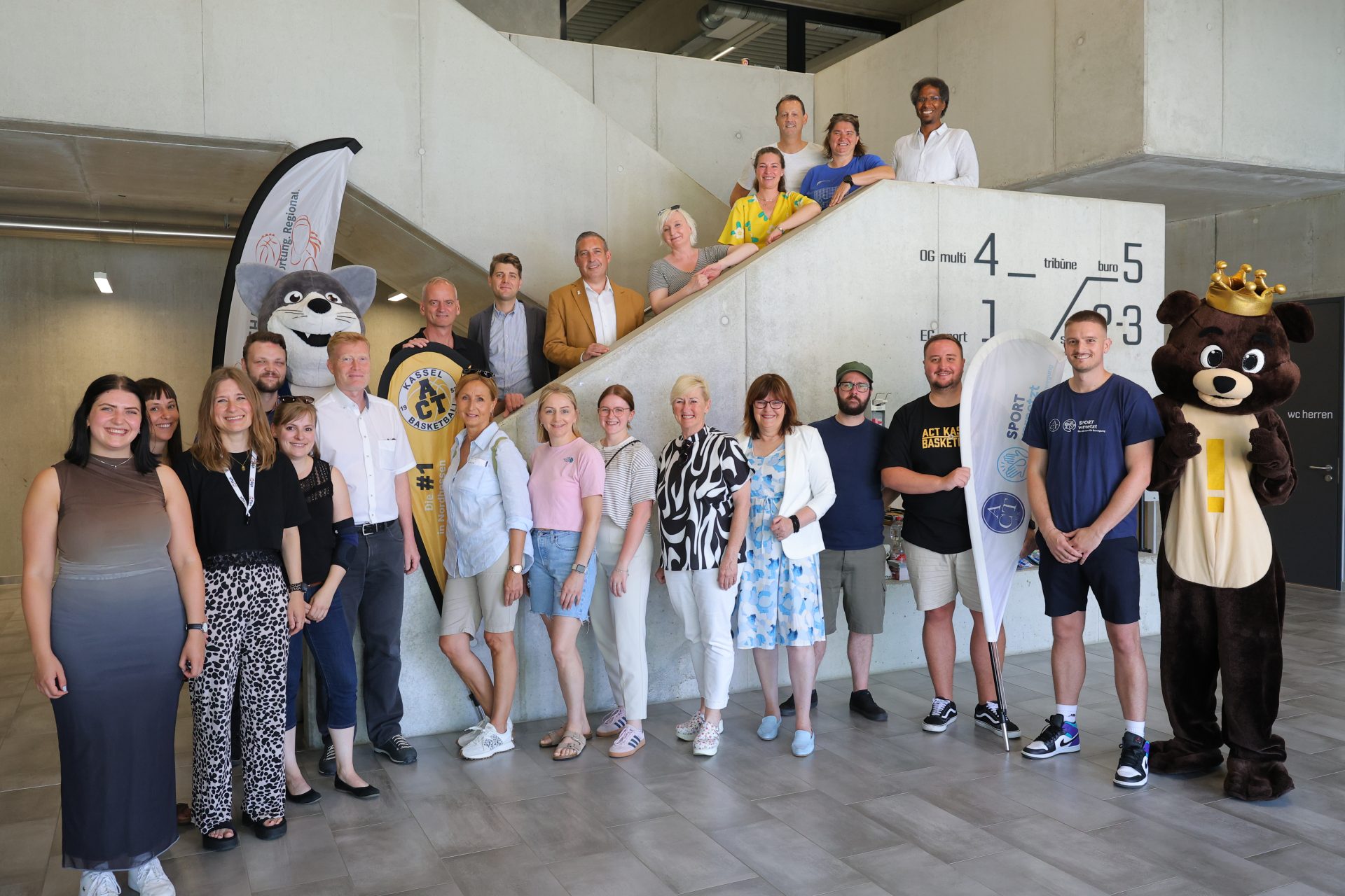 Gruppenfoto mit rund 25 Personen, zwei Maskottchen und zwei Beachflags in einem Treppenhaus, darunter Sascha Holstein, NHW-Regionalcenterleiter Kassel, Alexander Hauschild (Soziale Quartiersentwicklung NHW) und Kassels Sportdezernent Heiko Lehmkuhl