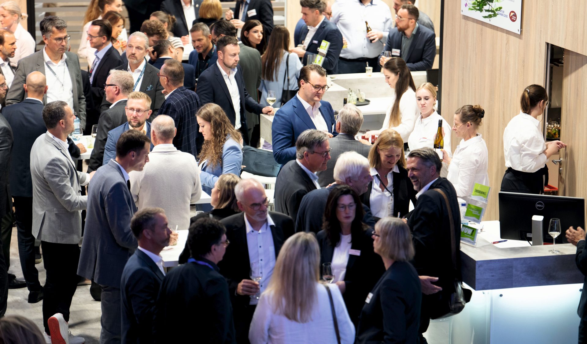 Viele Menschen, alle schick gekleidet, stehen an einem Messestand und unterhalten sich miteinander