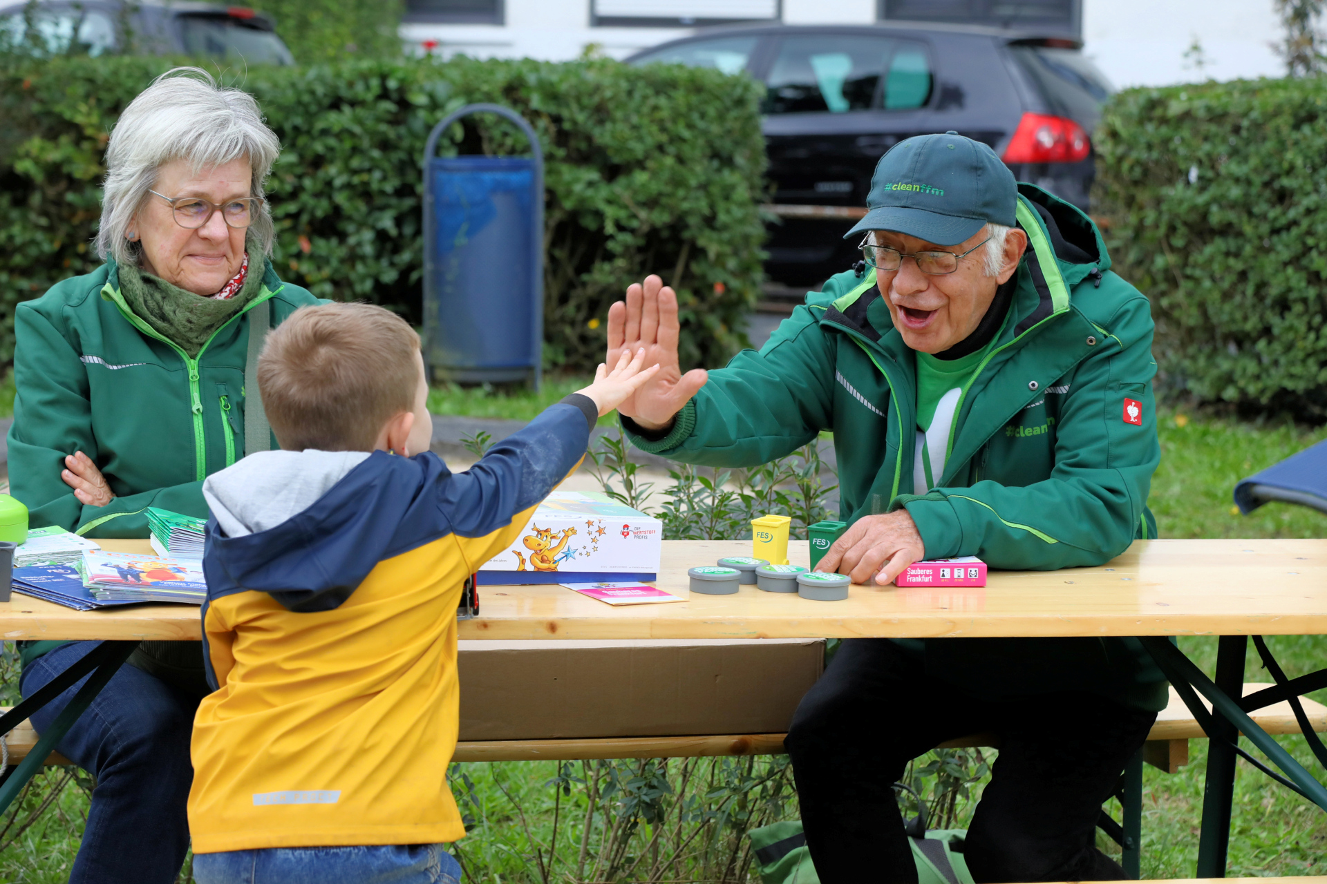 Ein älterer Mann und eine ältere Dame sitzen hinter einem Holztisch, der Mann gibt einem Jungen High Five