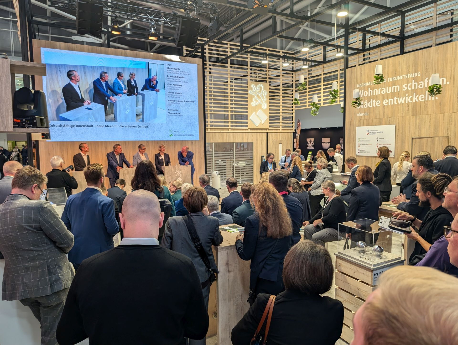 Eine große Zahl von Menschen hört interessiert anderen Menschen zu, die in einer Messehalle an Rednerpulten zum Thema Zukunftsfähige Innenstadt diskutieren.