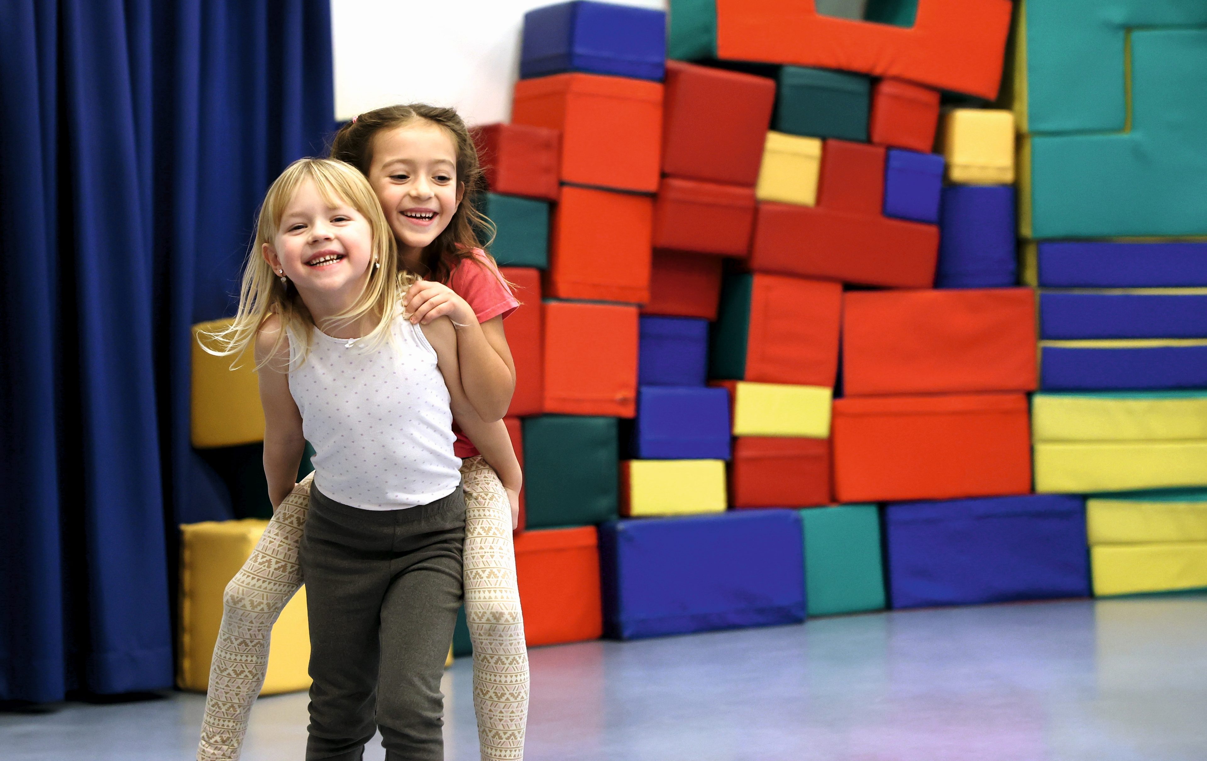 Ein Mädchen trägt ein anderes Mädchen in einer Turnhalle Huckepack. Beide strahlen. Im Hintergrund bunte Turnelemente.