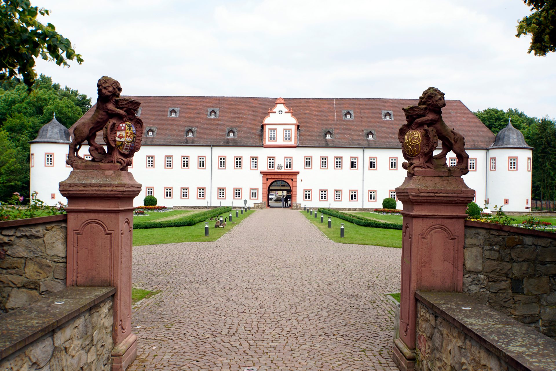 Blick auf ein schlossähnliches Gebäude mit einer gekiesten Auffahrt und zwei steinernen Figuren auf zwei Sockeln im Vordergrund