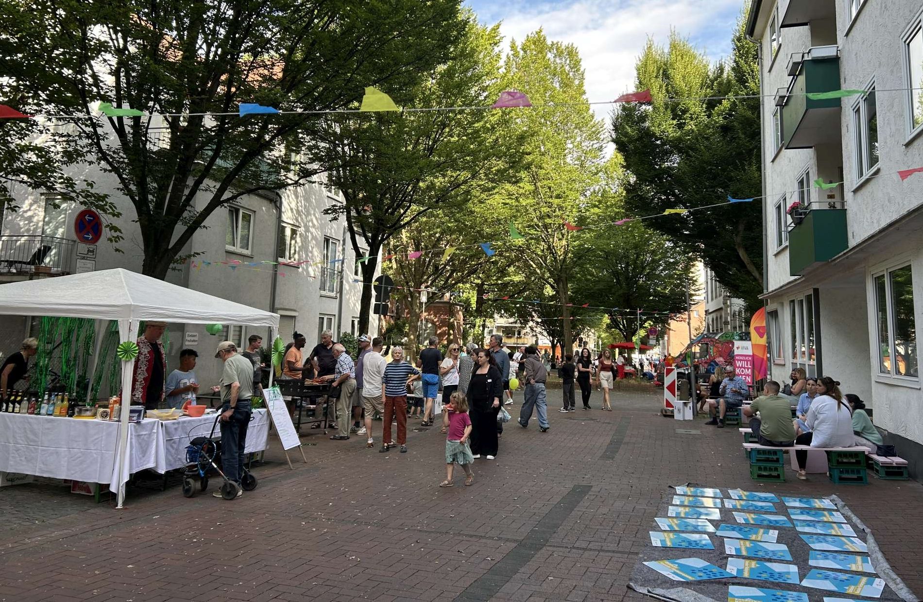 Mit Wimpeln geschmückte Straße, Menschen, die auf Bänken sitzen, vereinzelte Stände