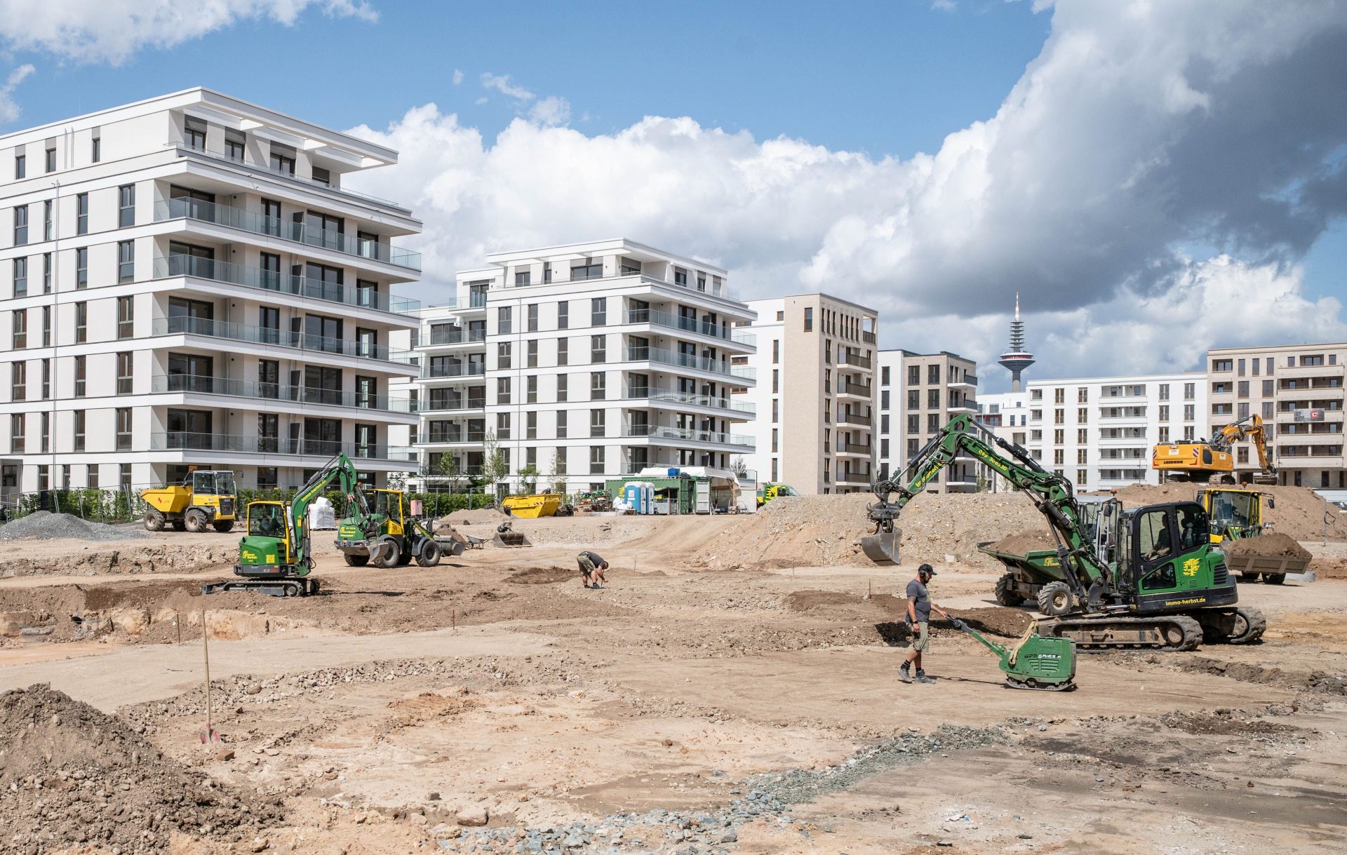 Eine Baustelle mit weitgehend fertiggestellten Neubauten im Hintergrund, im Vordergrund eine riesige Baugrube, Baustellenfahrzeuge und Bauarbeiter.