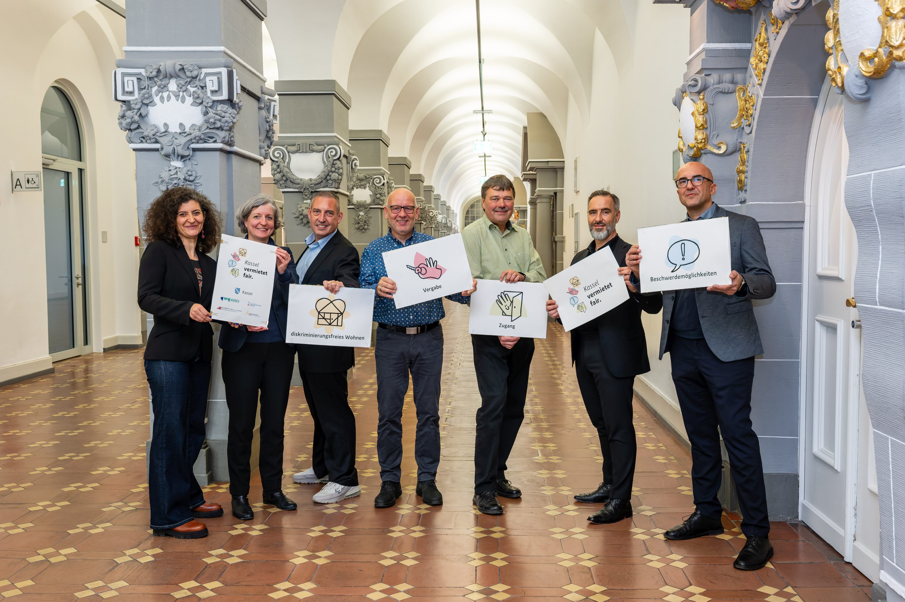 Mitglieder der Initiative Kassel vermietet fair. Jeder hält eine Tafel mit einzelnen Elementen des Leitbilds in die Höhe (v.l.n.r.): Teslihan Ayalp (Leiterin Amt für Chancengleichheit), Stadtbaurätin Simone Fedderke, Sascha Holstein (NHW, Leiter Regionalcenter Kassel), Sven Hansen (Bauverein 1894), Michael Fuchs (Studierendenwerk), Uwe Gabriel (GWG), Evren Özgüvenc (Antidiskriminierungsbeauftragter).