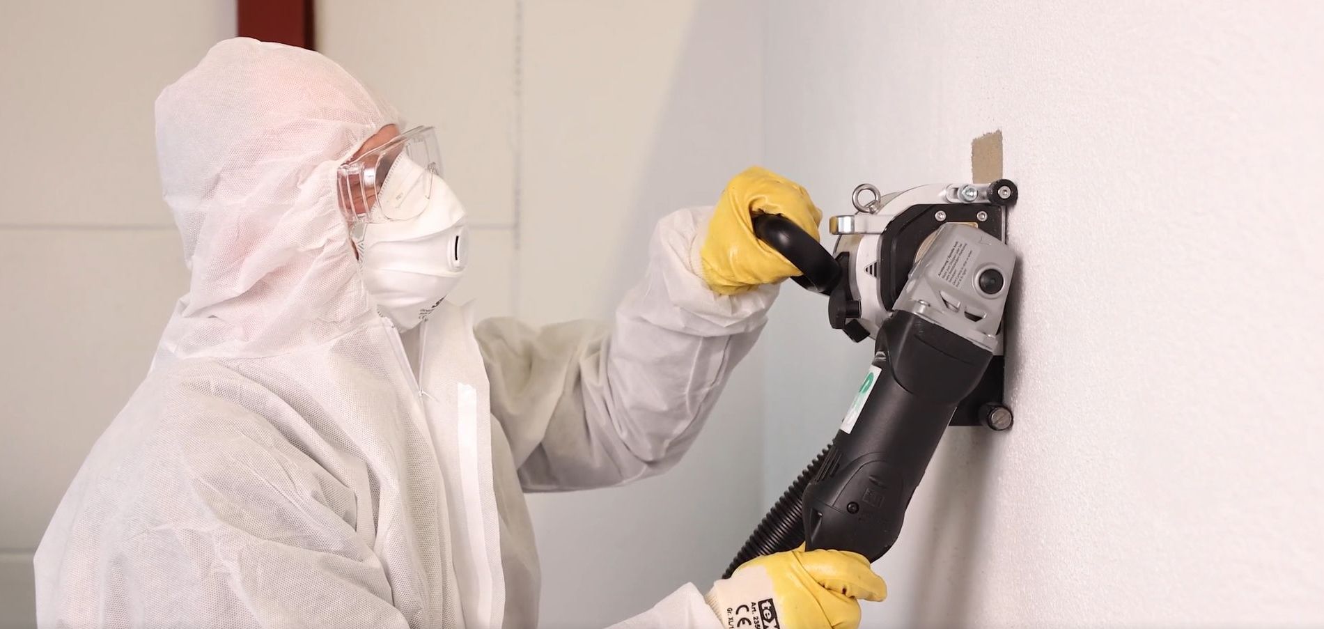 Mann in Schutzanzug entfernt maschinell Asbest von der Wand.