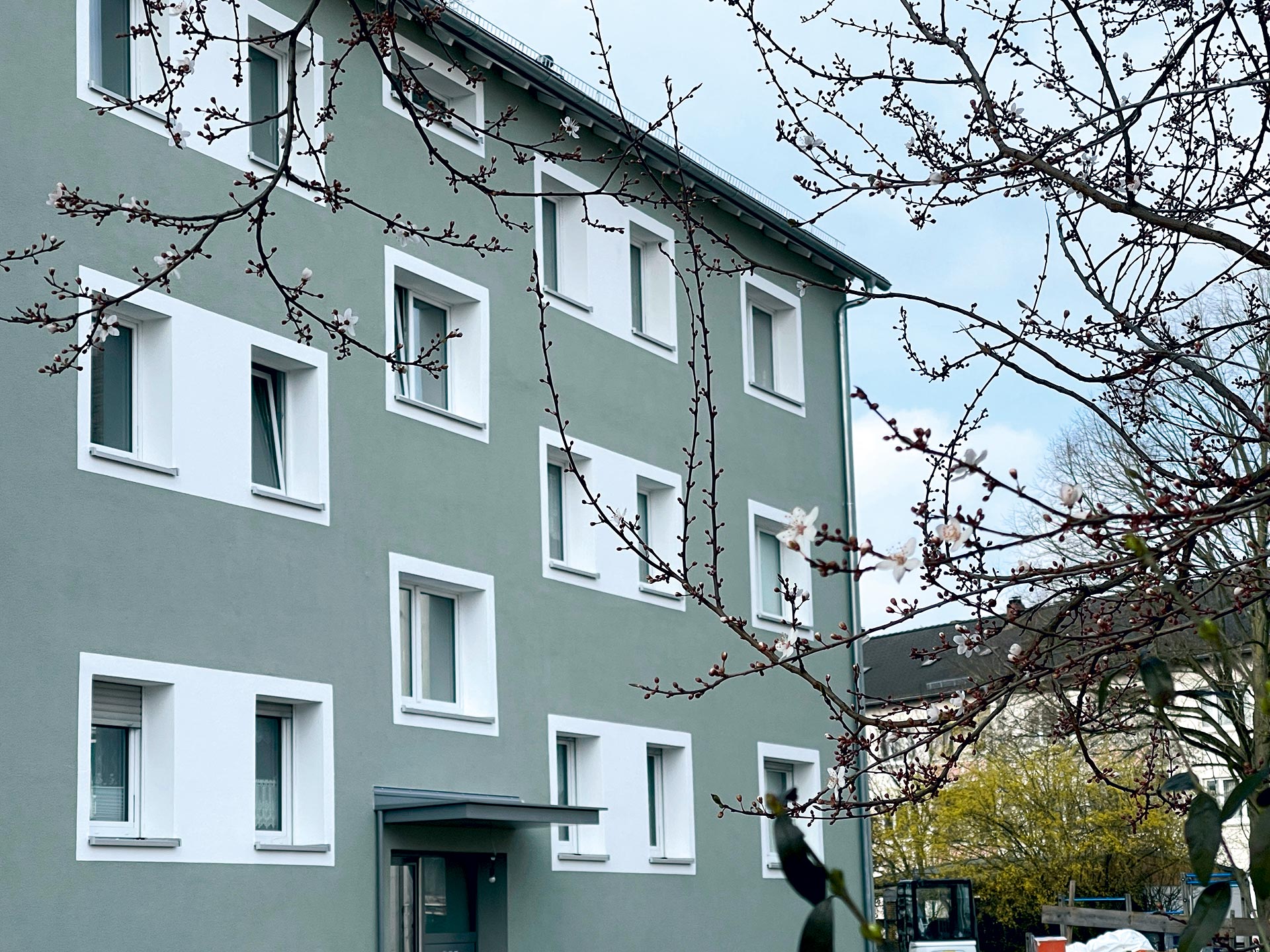 Ein fertig saniertes, grau-blaues Wohnhaus mit weißen Fenstern, fotografiert durch die Äste eines kahlen Baumes.