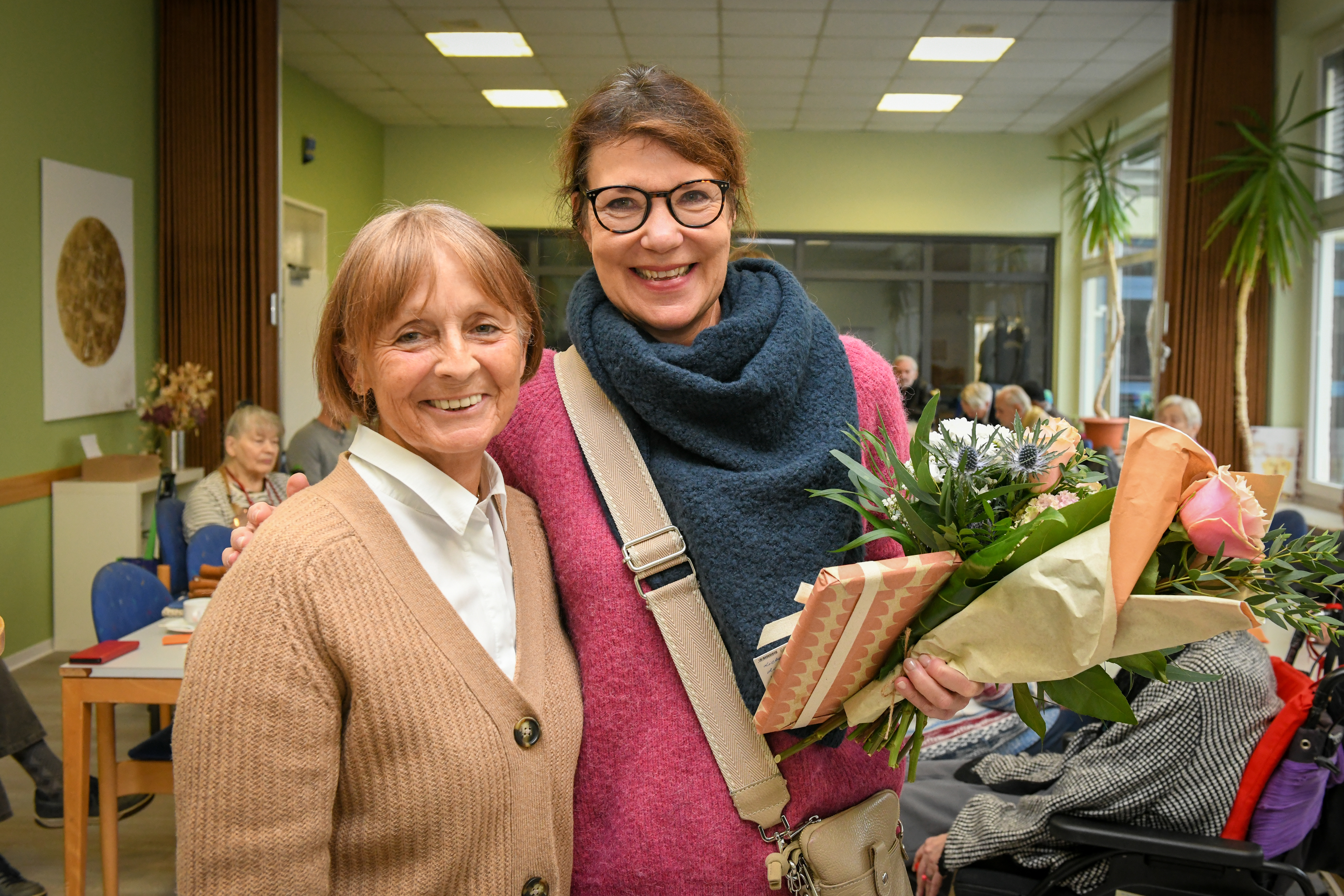 Mitarbeiterin von NHW und Leiterin vom Volksbildungswerkt steht mit einem Blumenstrauß daneben. Beide lachen in die Kamera.