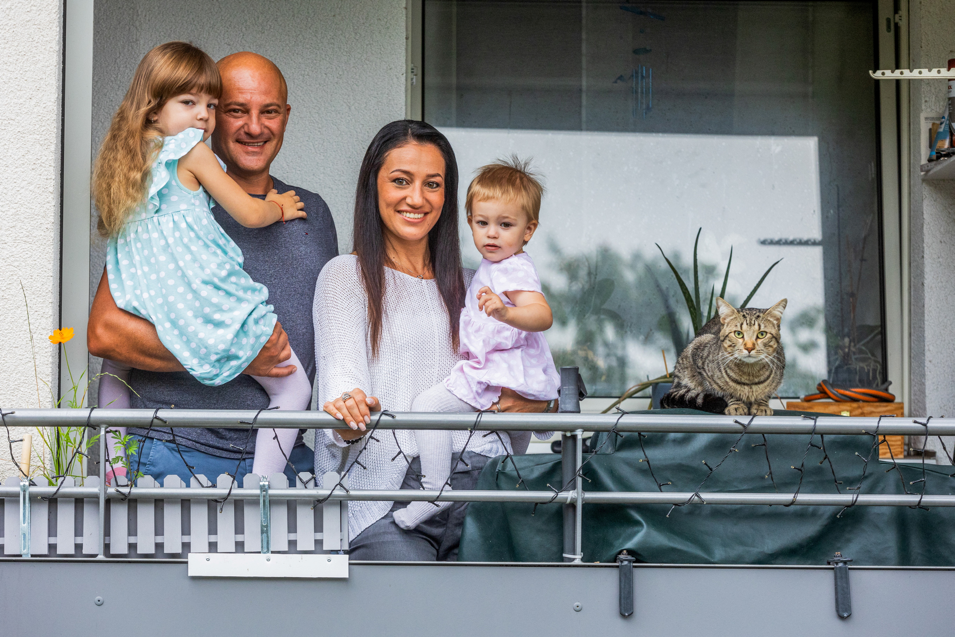 Familie steht an einem Balkon und schaut in die Kamera. Mann hält kleine Tochter im Arm, die Frau hält ein Baby im Arm.