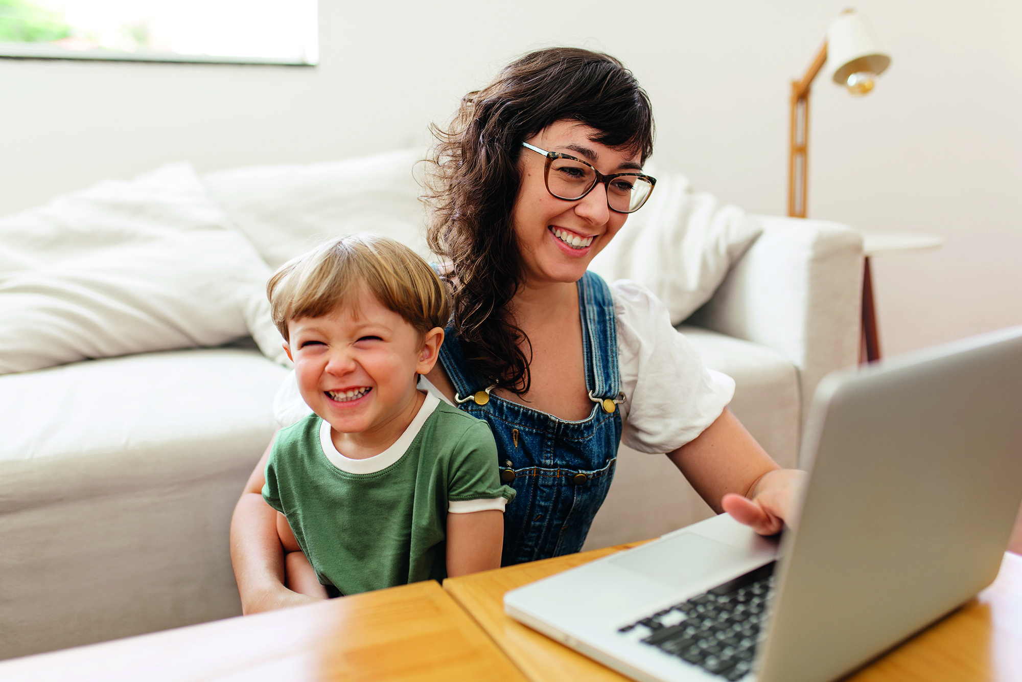 Frau mit Brille sitzt mit einem Kleinkind vor einem Laptop. Kleiner Junge lacht in die Kamera, Frau schaut in den Laptop.