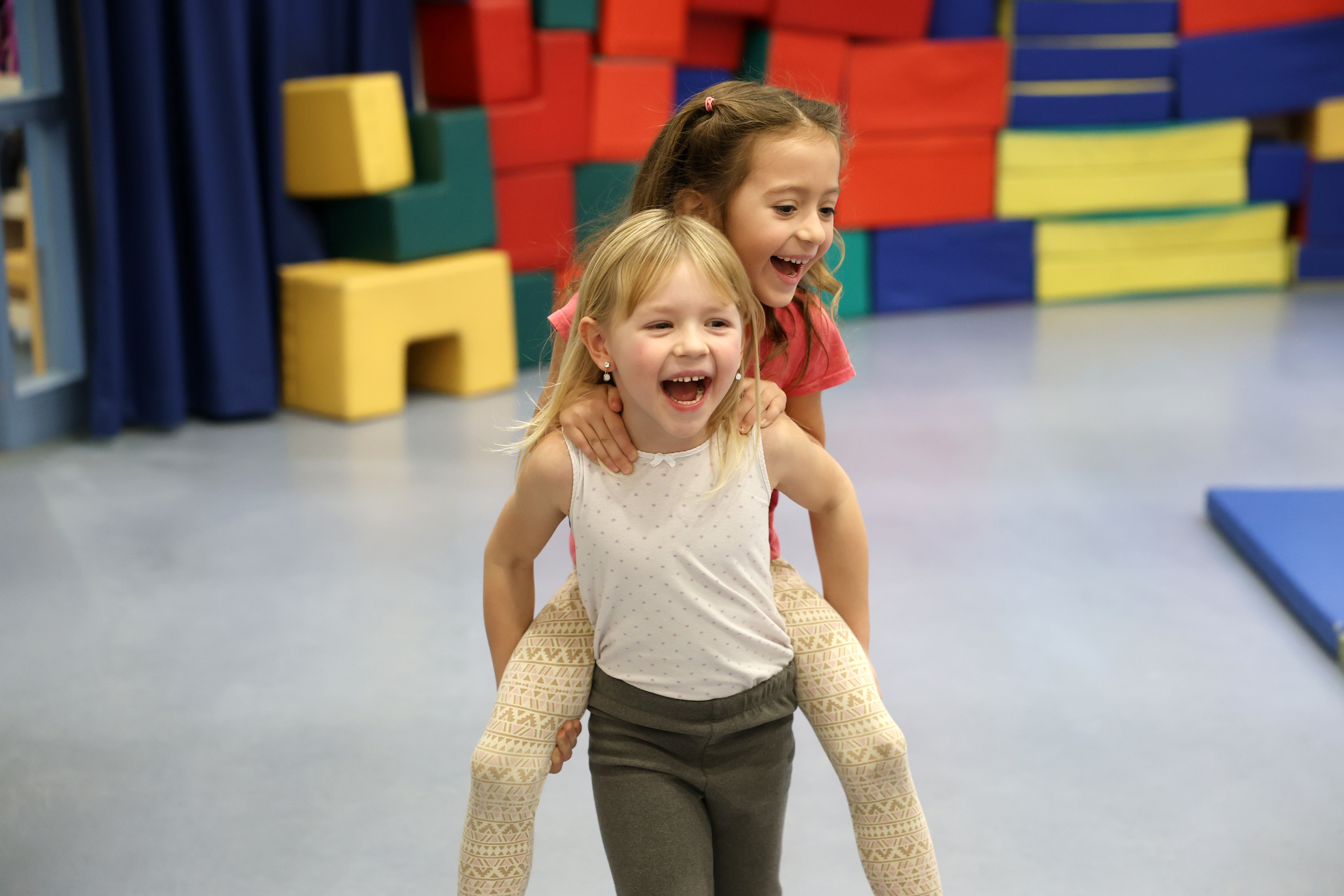 Zwei Kinder laufen in einer Sporthalle. Im Hintergrund sind Matten zu sehen. Ein Kind trägt das andere Huckepack. Kinder lachen.