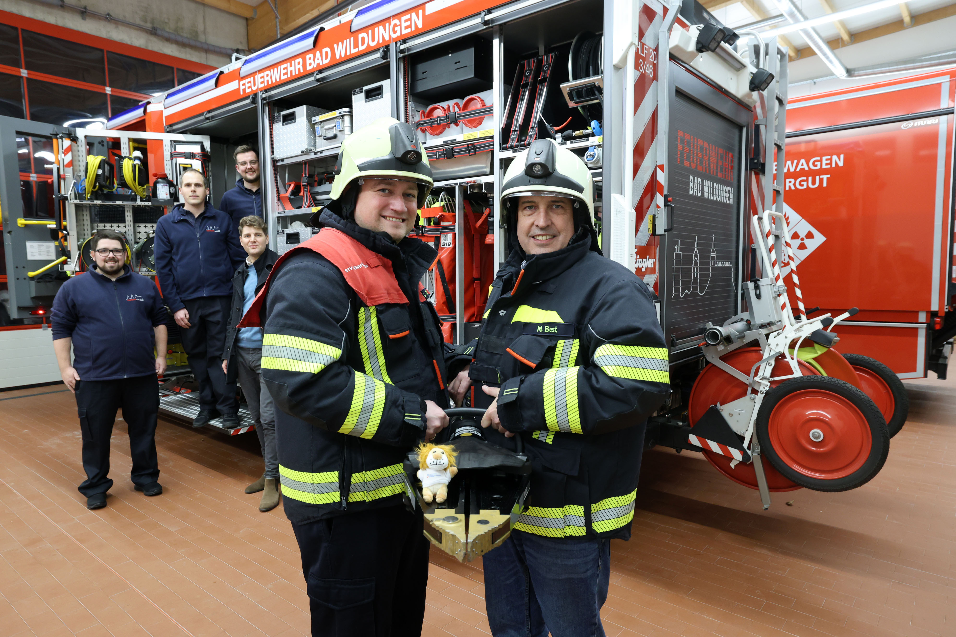 Im Vordergrund sind zwei Feuerwehrmänner mit Ausrüstung. Im Hintergrund steht ein Löschfahrzeug und weitere Personen steigen aus dem Feuerwehrauto aus.