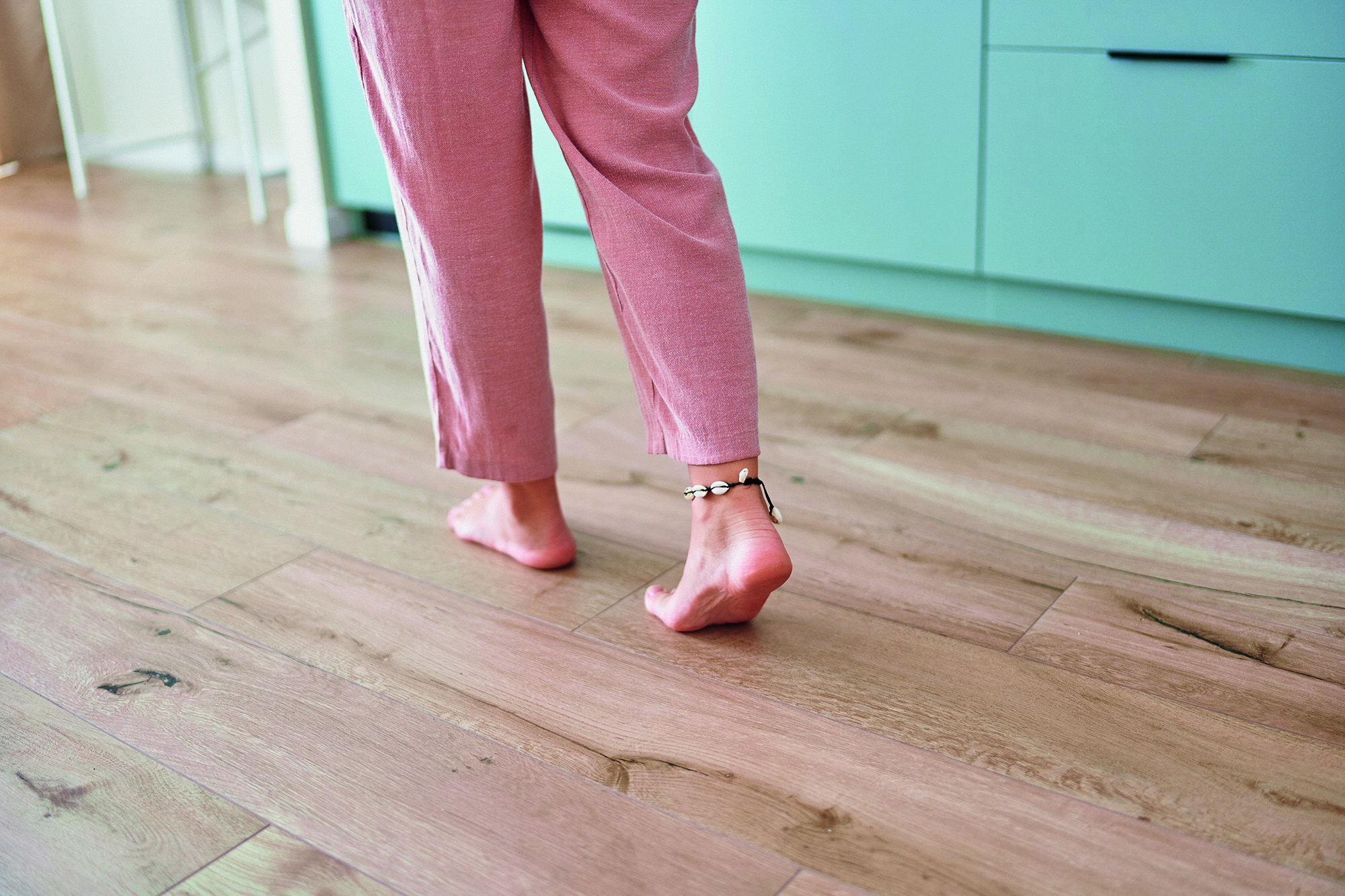 Frau läuft barfuß über einen Parkettboden. Oberkörper ist abgeschnitten. Fokus auf den Boden.