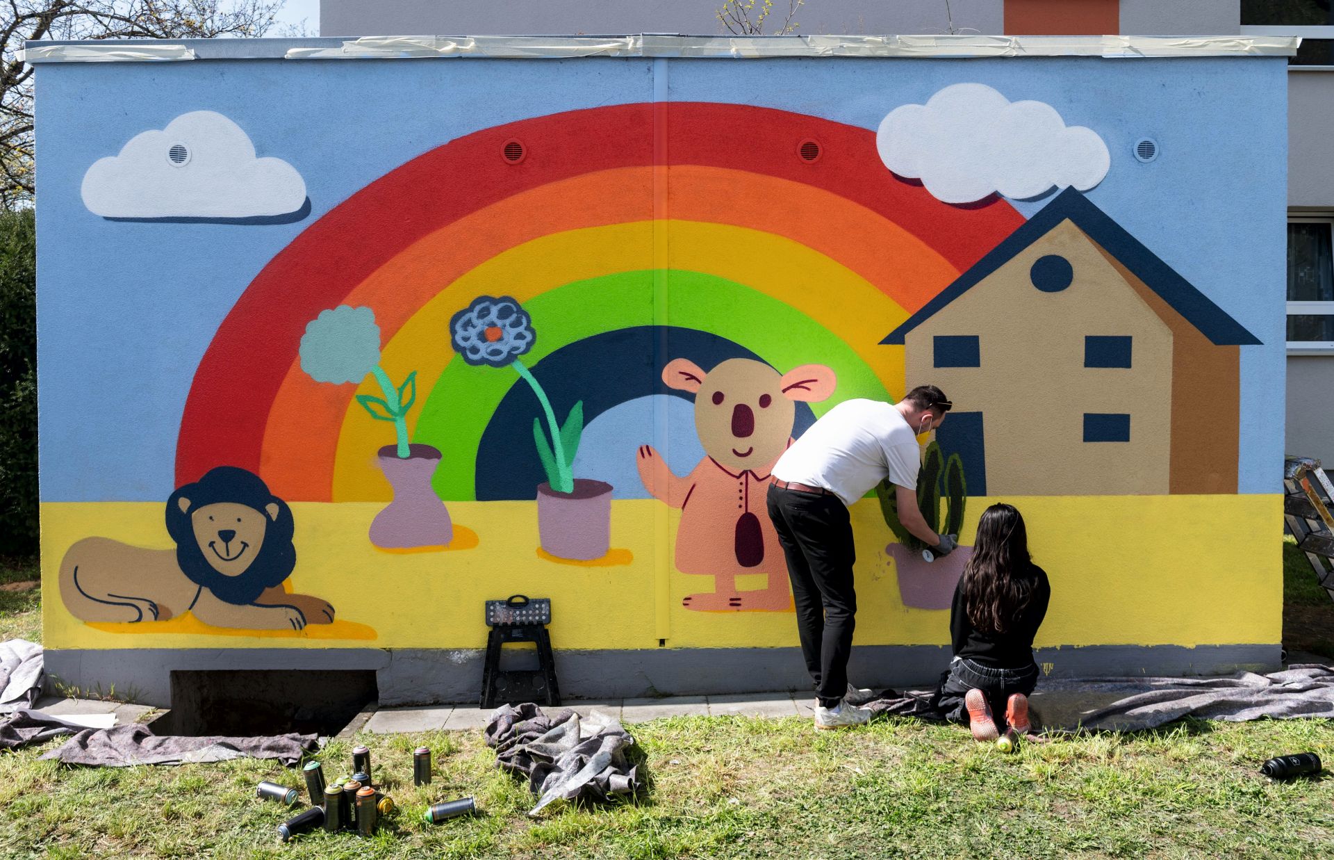 Wand mit einem Graffiti. Regenbogen mit Haus und Blumen im Topf, ein Löwe. Mädchen sprüht und ein Anleiter steht dabei.
