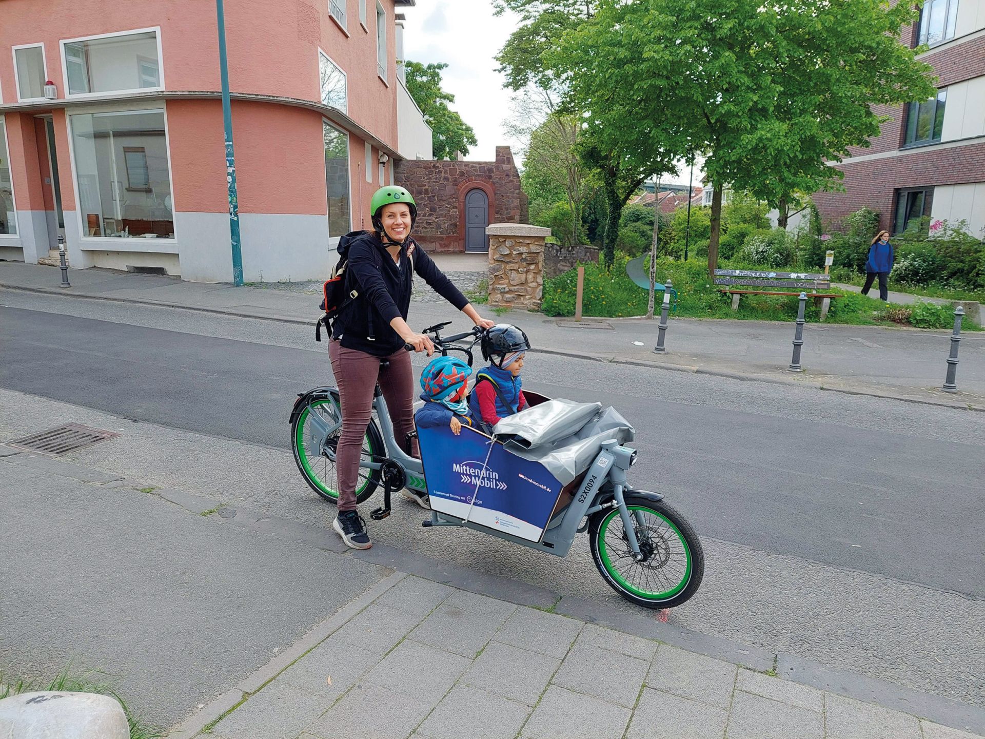 Frau mit zwei Kindern in einem Lastenrad. Alle tragen Helme.
