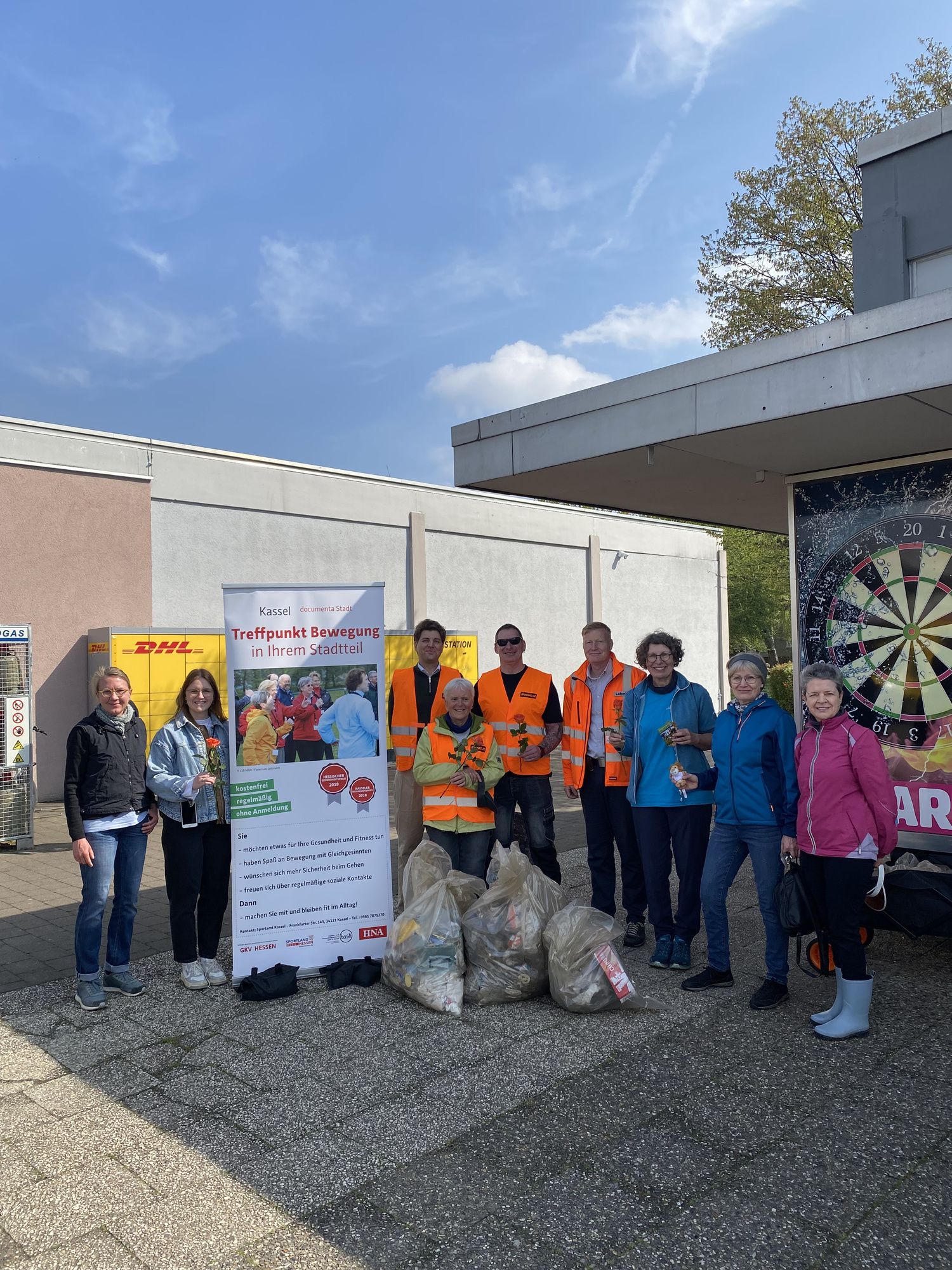 Gruppe von Menschen mit gesammelten Müllbeuteln im Vordergrund. Aufsteller "Treffpunkt Bewegt"