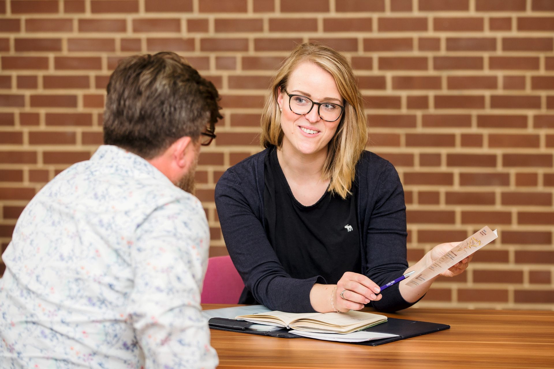 Frau sitzt vor einer rot gekachelten Wand und berät eine Person. Person sitzt mit dem Rücken zur Kamera.
