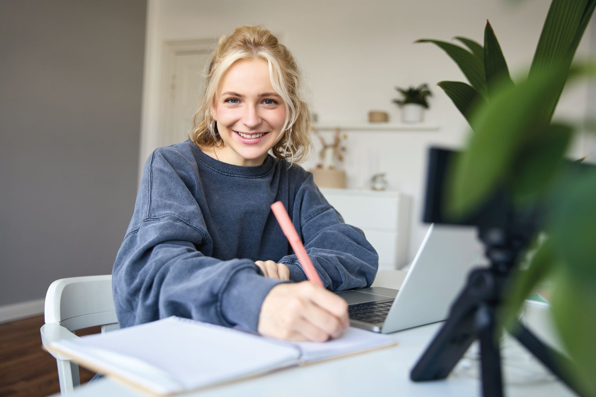 Mädchen sitzt an einem Laptop und schreibt etwas auf einen Block neben dem Laptop. Mädchen sieht in die Kamera und lacht.