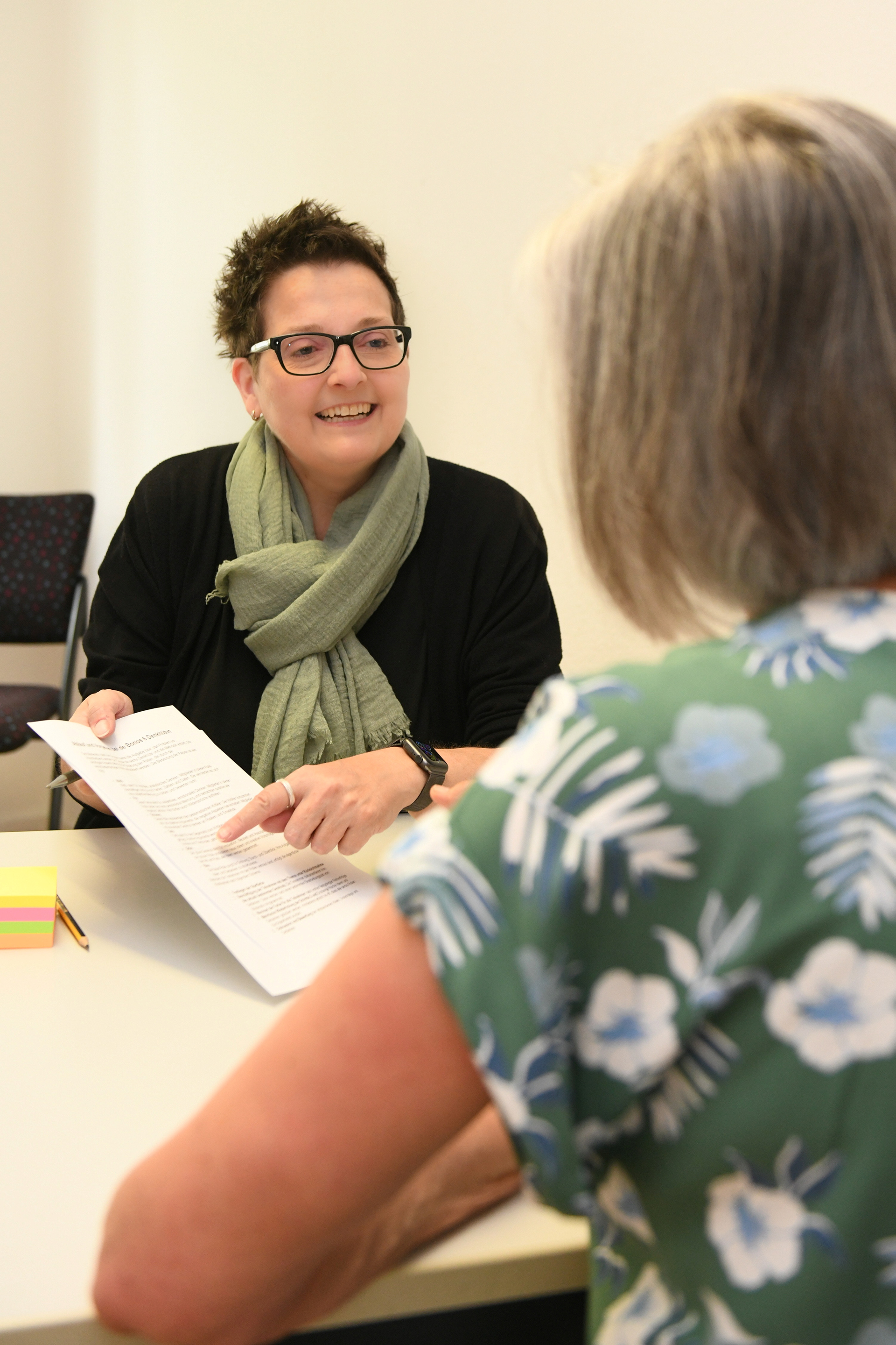 Frau Sitz an einem Schreibtisch mit einem Blatt in der Hand und berät eine andere Frau, die mit dem Rücken zur Kamera sitzt