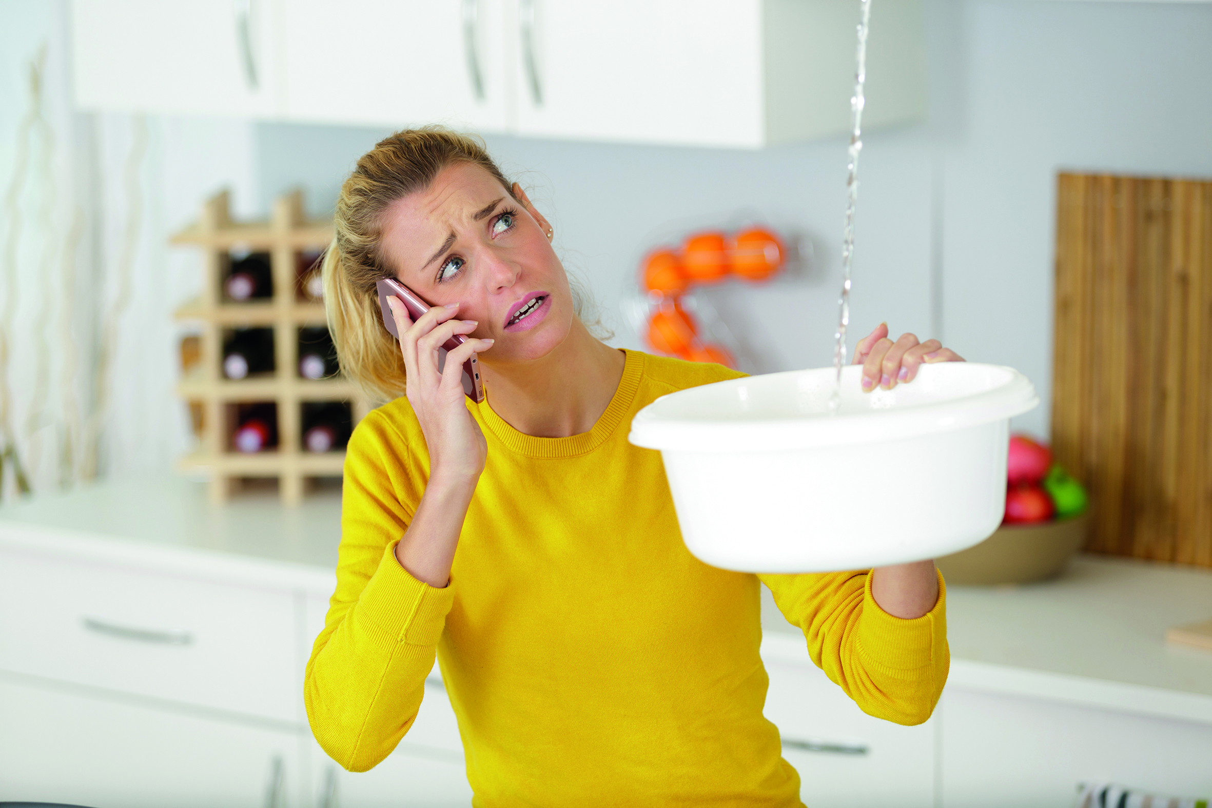 Frau hält eine Schüssel in der Hand und fängt tropfendes Wasser von der Decke auf. Hat das Handy am Ohr und ruft jemanden an