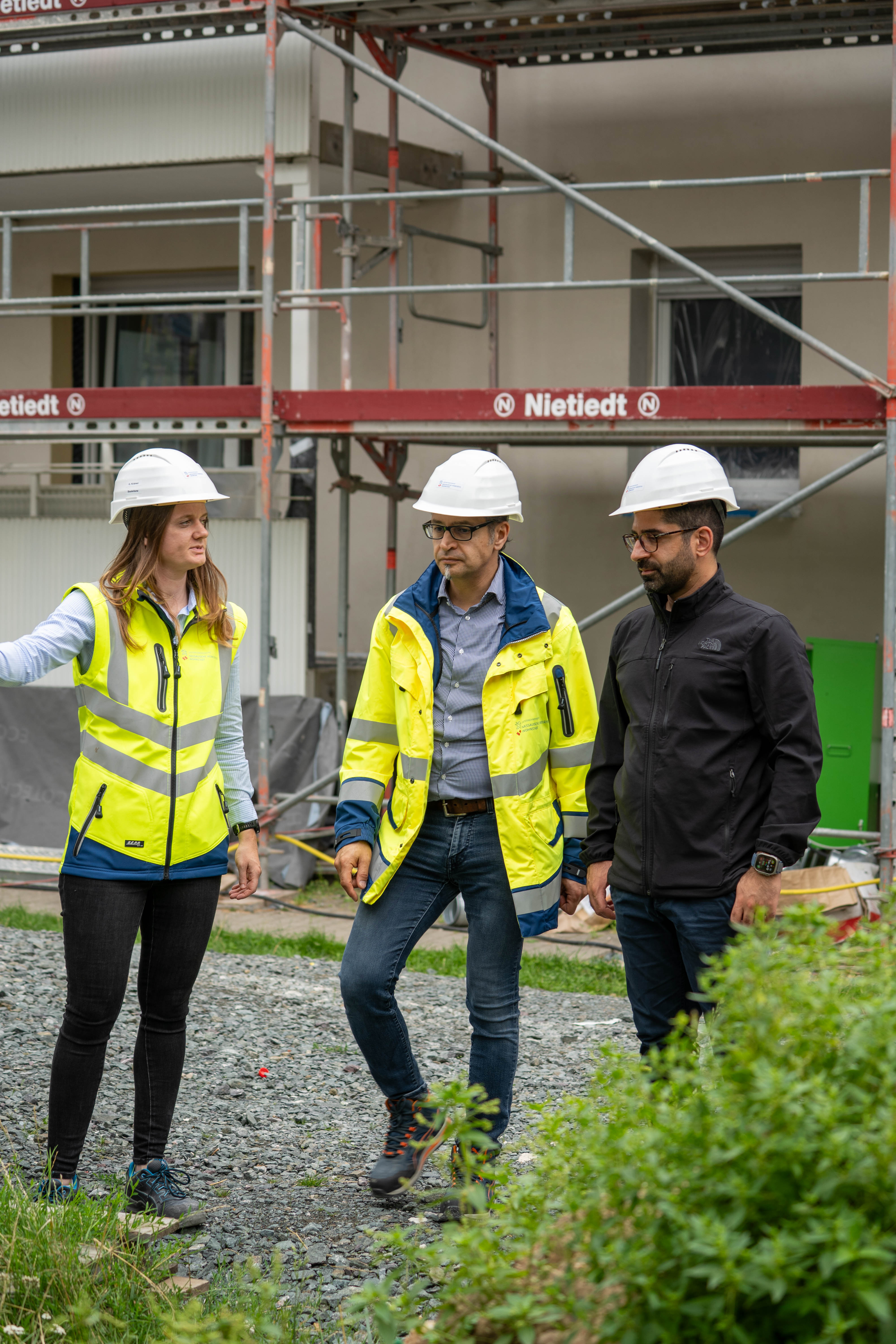 Frau und Mann mit Warnweste und weißen Bauhelm an. Dritte Person ohne Warnweste, schwarze Kleidung mit weißen Helm.